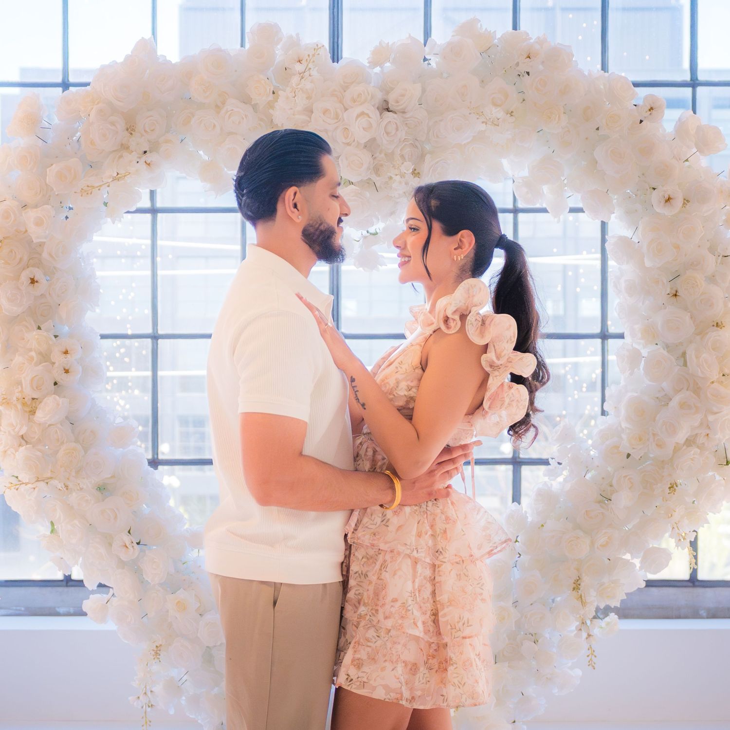 Couple smiling at each other in front of a beautiful white floral heart arch arrangement.