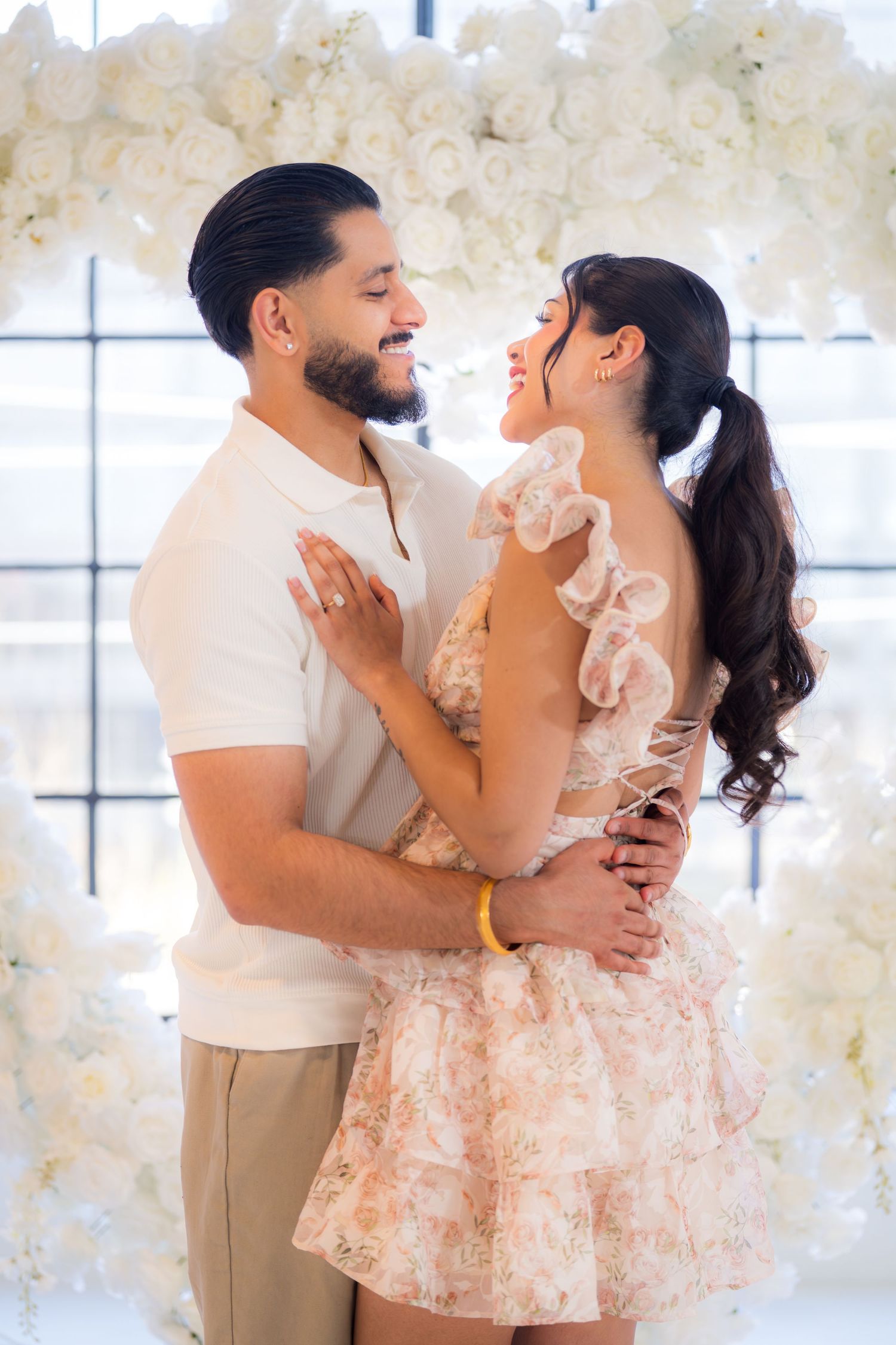 Romantic couple embracing and smiling at each other in front of white rose floral arch backdrop.