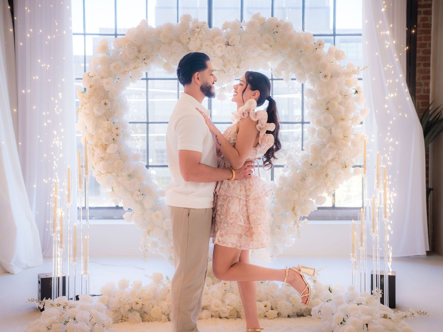 Romantic couple standing face to face in front of large white floral heart arch with candles.
