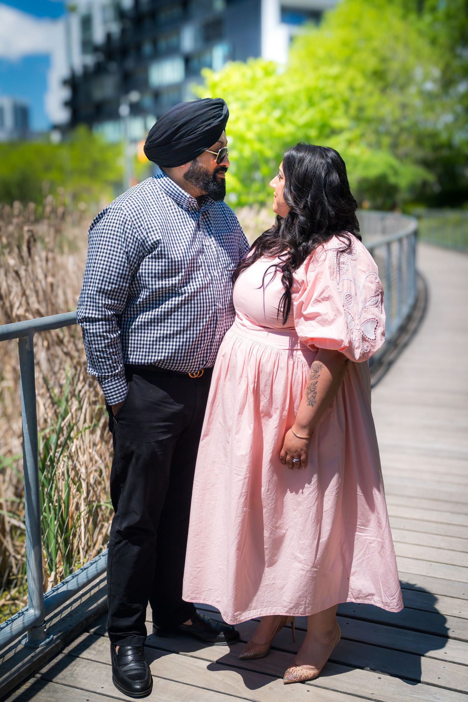 A Sikh couple shares a loving gaze on a wooden boardwalk surrounded by lush green trees.
