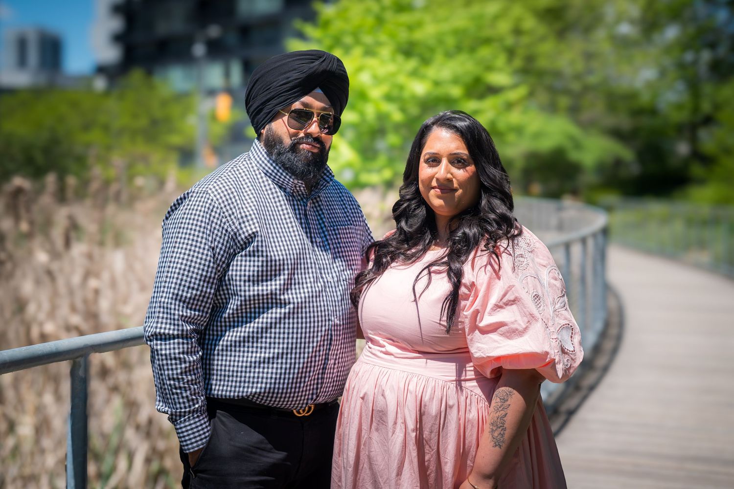 Sikh man in blue turban and woman in pink dress pose on a sunny outdoor walkway surrounded by greenery.