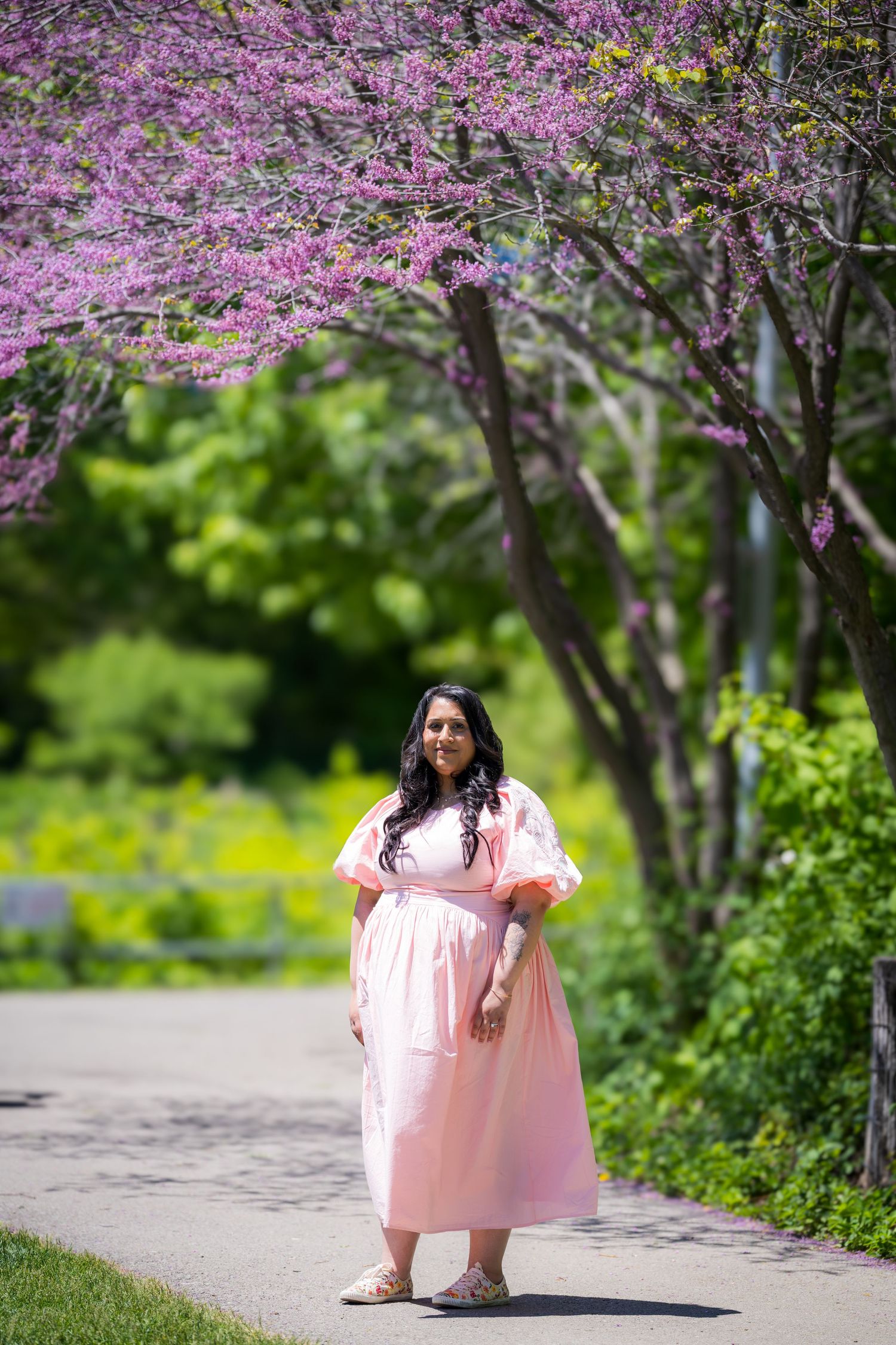 A pregnant woman in a flowing pink dress stands on a path surrounded by blooming purple redbud trees.