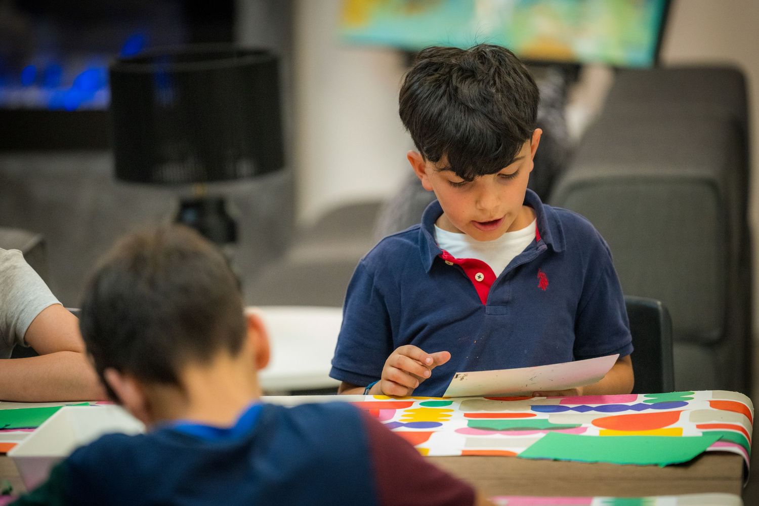 Young boy in blue polo shirt focused on a colorful craft activity at a classroom table.