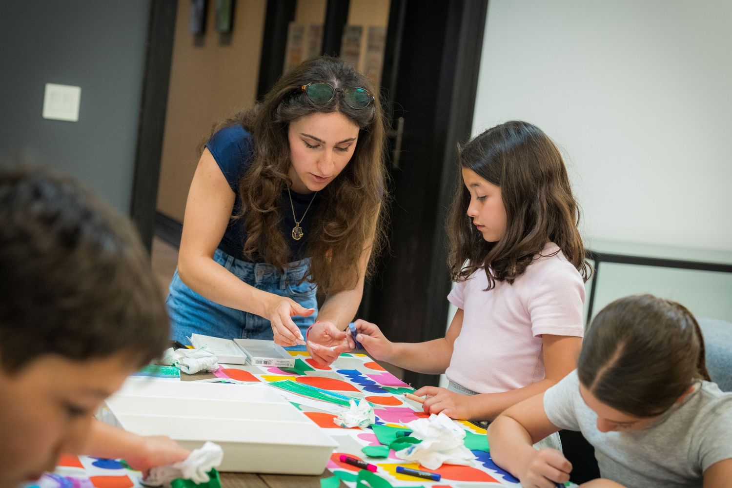 A teacher assists children with a vibrant paper collage art activity at a classroom table.