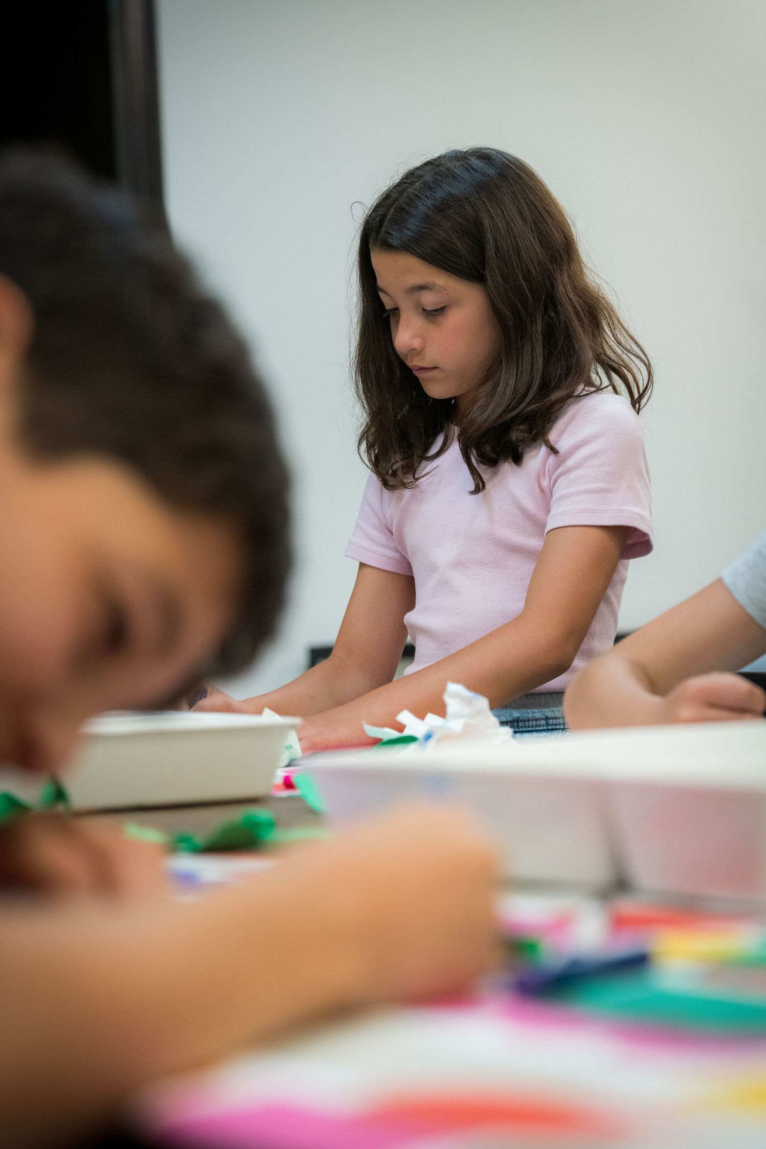Girl in pink shirt concentrates on colorful paper collage while boy works in foreground.