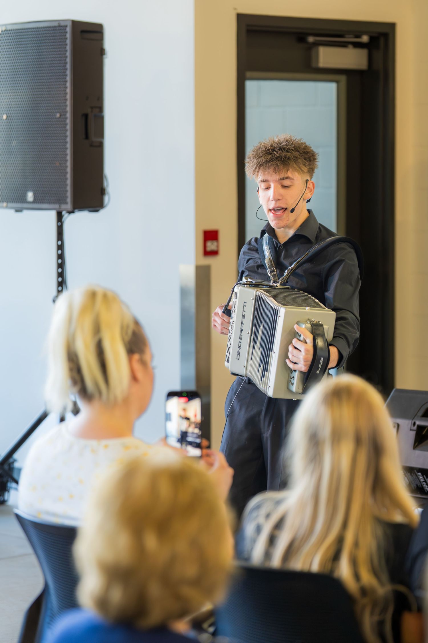 Young curly-haired musician singing and playing accordion for seated audience members.