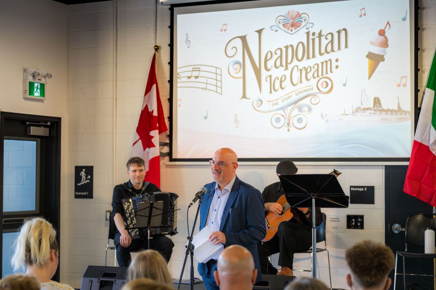 Speaker presenting Neapolitan Ice Cream show with musicians and Canadian flag at indoor event.