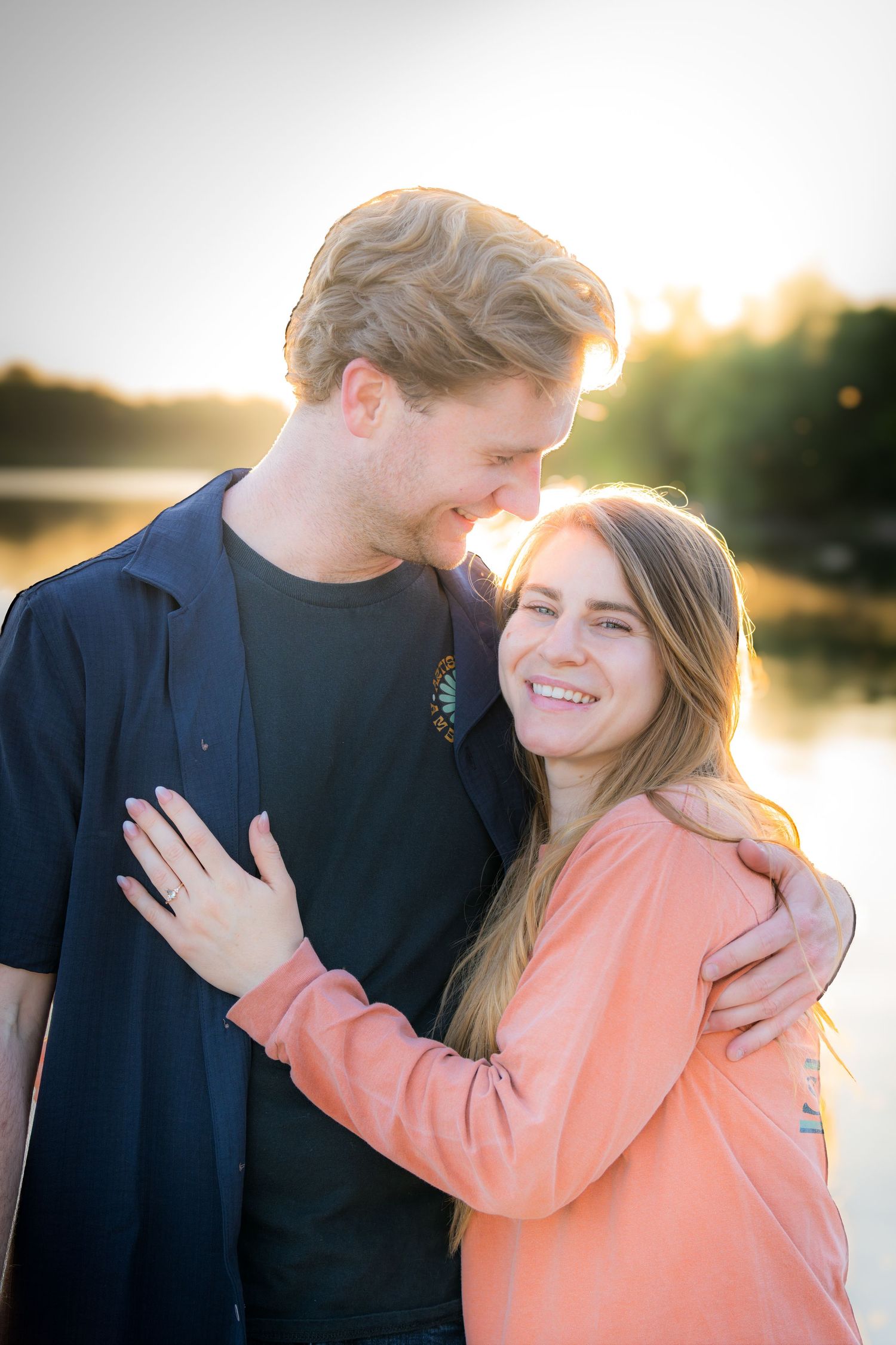 Couple embracing and smiling at sunset by a lake, man in navy shirt hugging woman in pink hoodie.