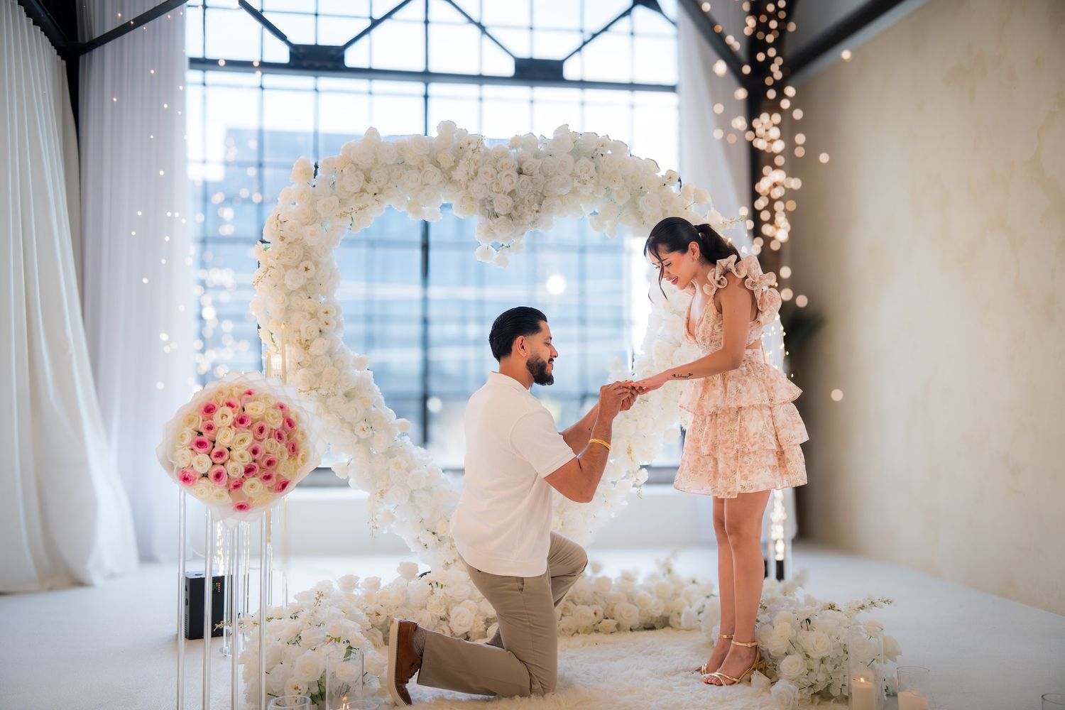 Man proposes to woman on one knee under a white floral heart arch in an elegant decorated indoor venue.