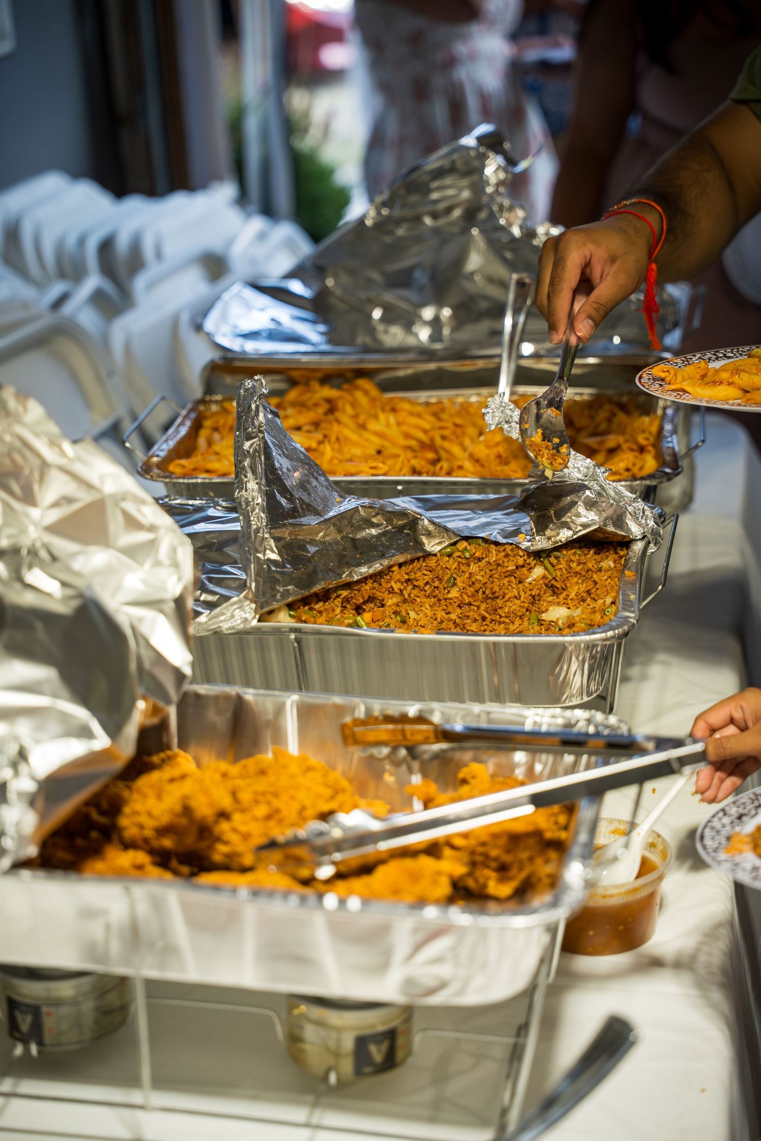 Buffet setup with multiple trays of orange-colored rice or pasta dishes being served with metal tongs.