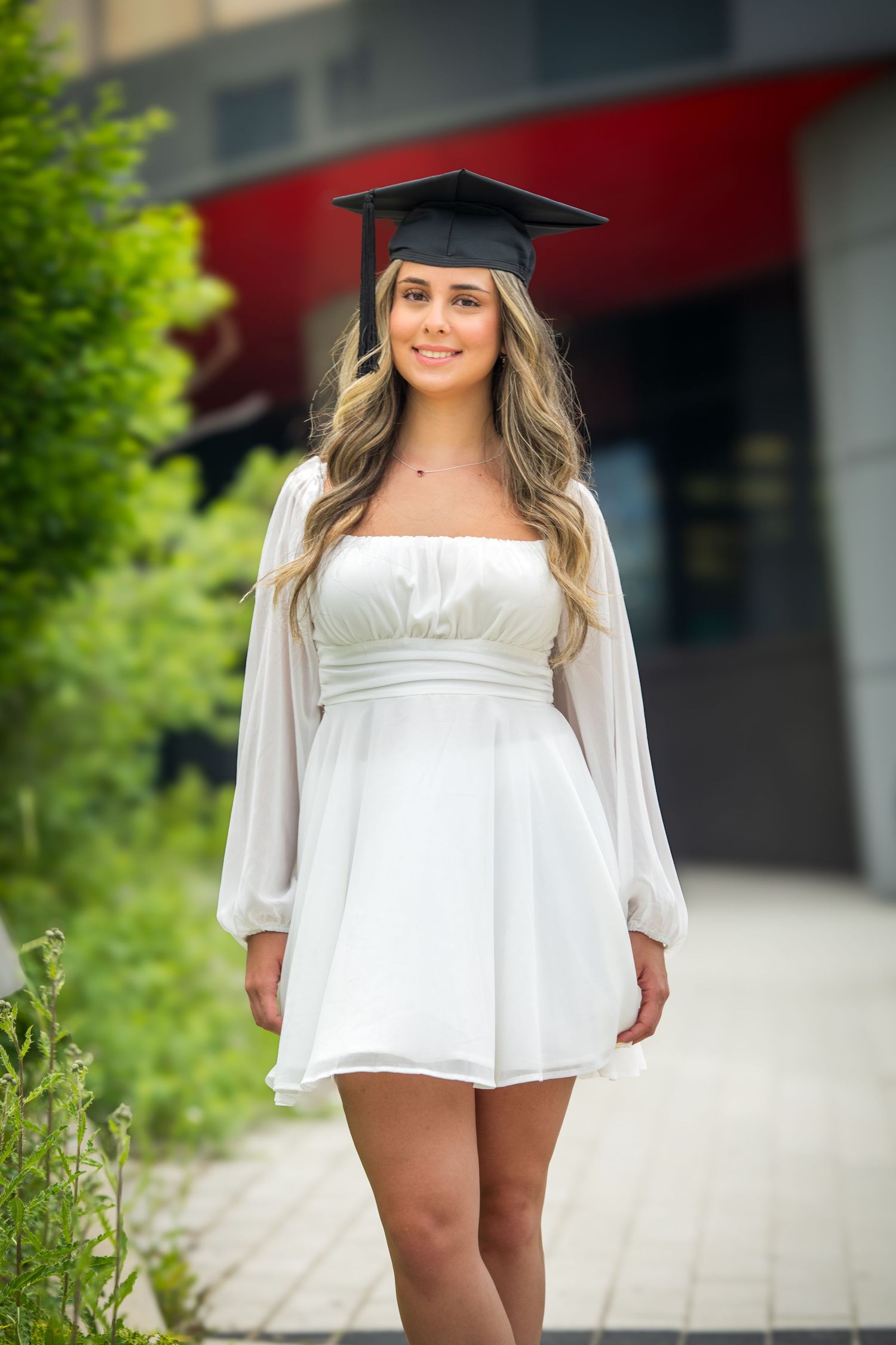Graduate in short white dress and cap poses outdoors near red building.