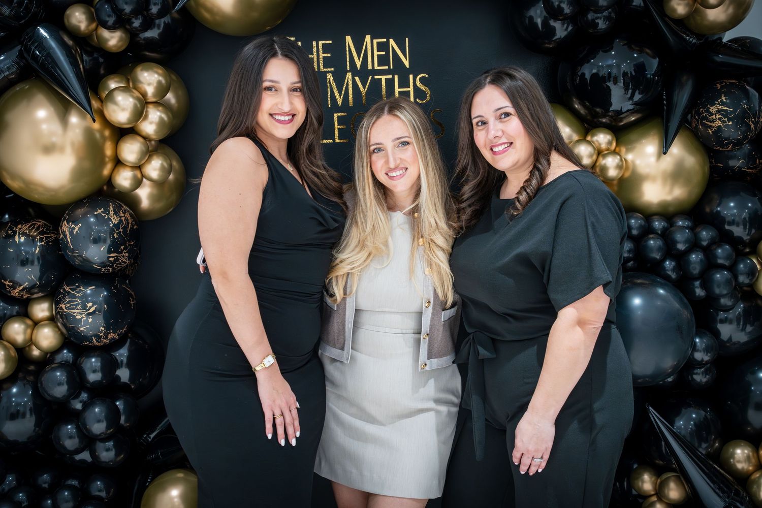 Three women posing together at event with gold balloons and black backdrop.