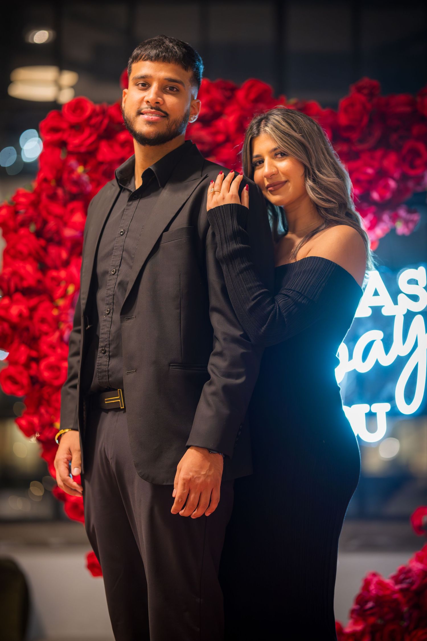 An elegantly dressed couple posing in front of a romantic red rose backdrop with neon signage.