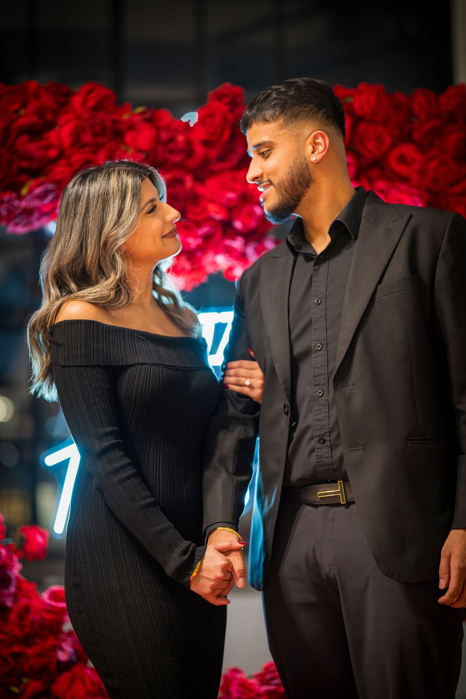 Romantic couple shares intimate moment in front of red floral wall with neon lighting.