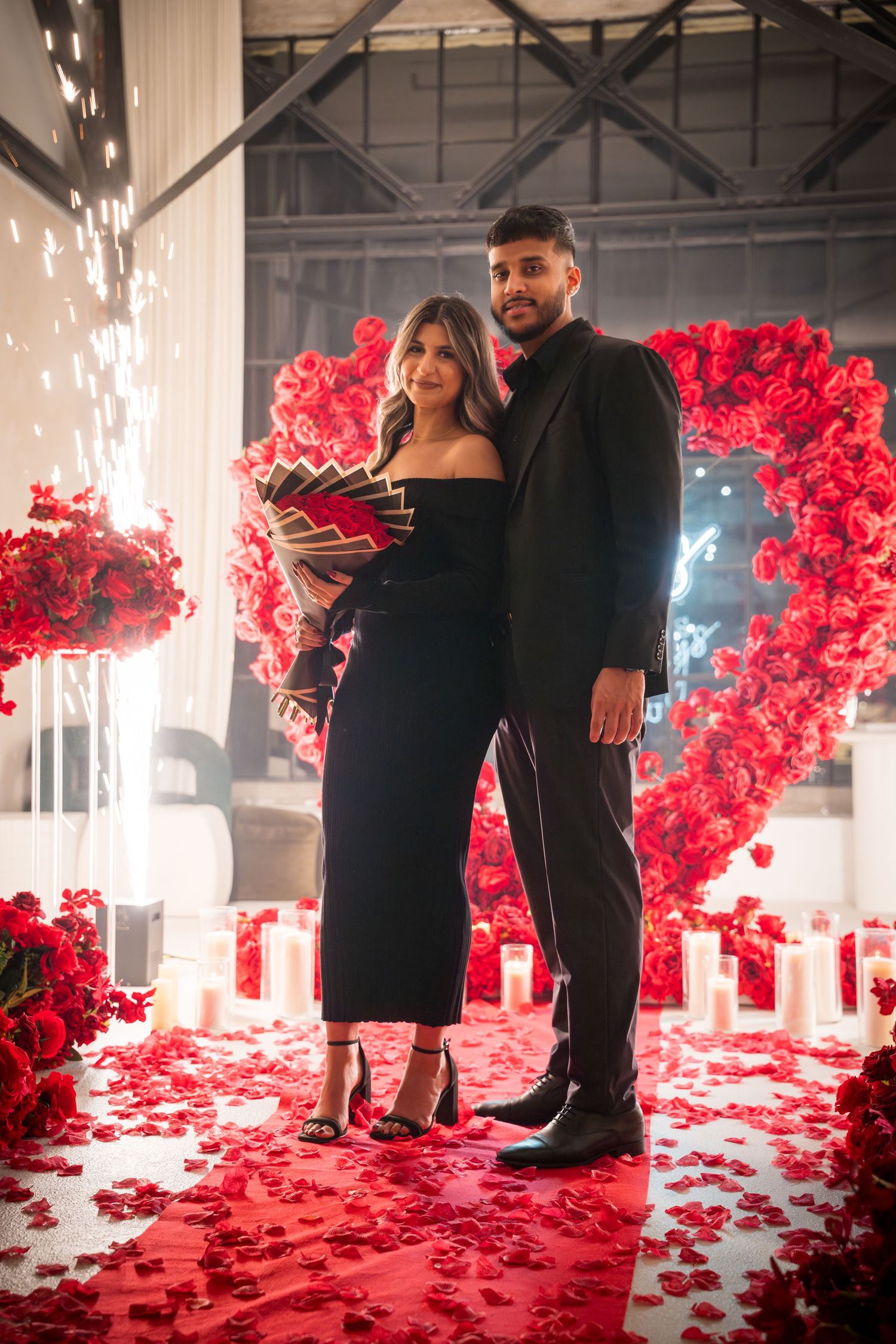 Couple standing in romantic setting with red roses, sparklers and neon lighting.
