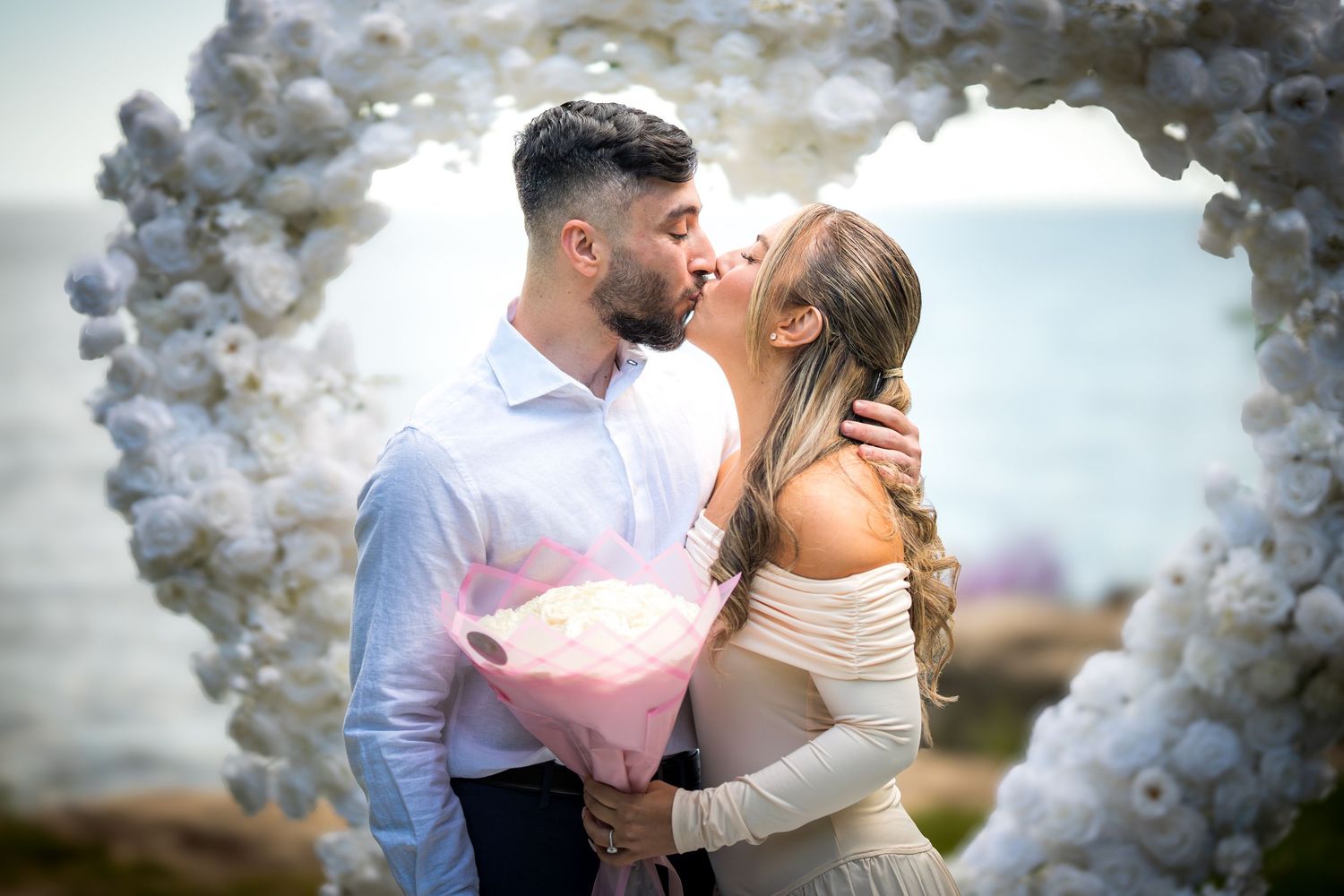 A romantic proposal moment under a white balloon arch by the ocean with a pink cotton candy bouquet.