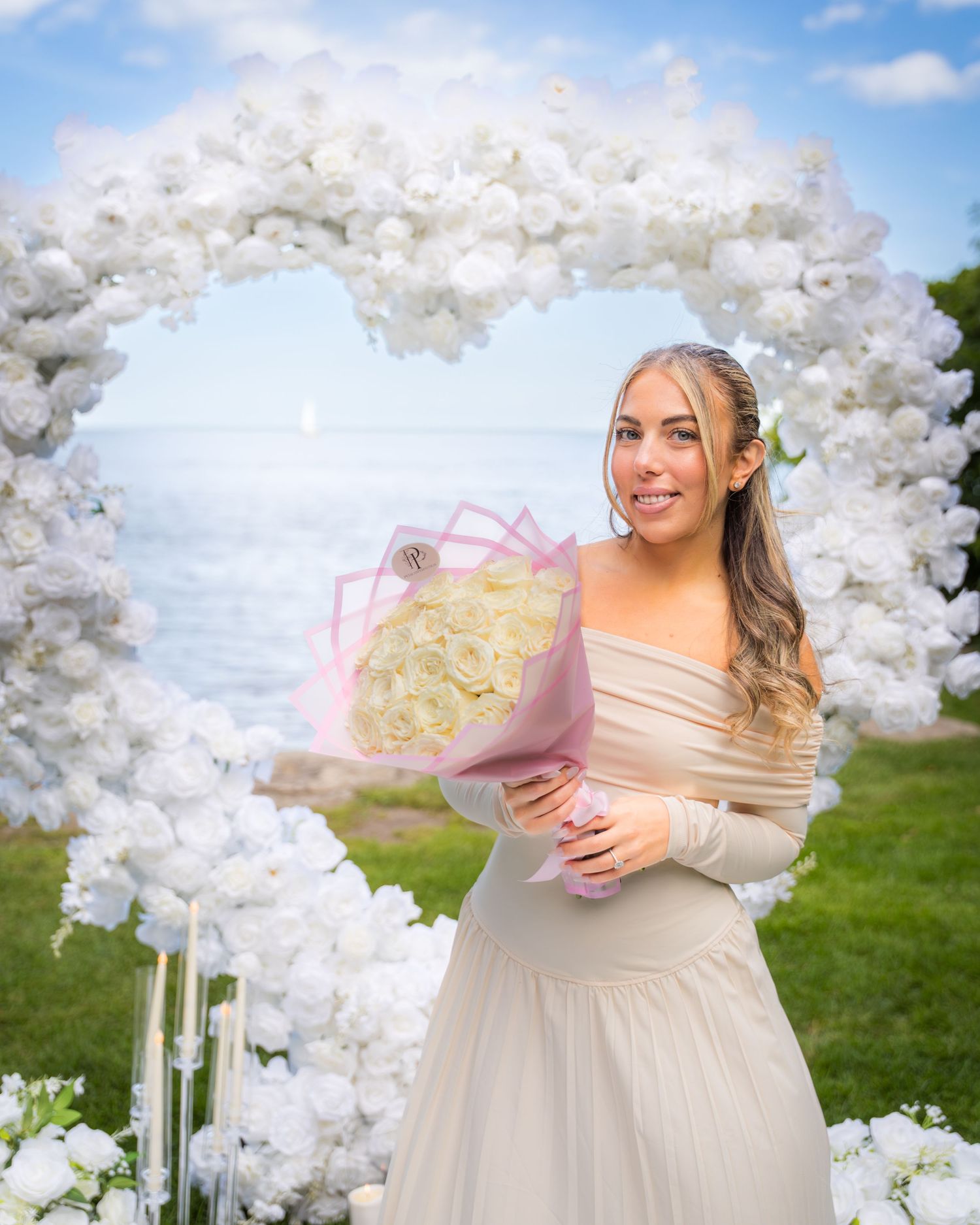 An elegant proposal setting with white floral arch and pink bouquet by the ocean.