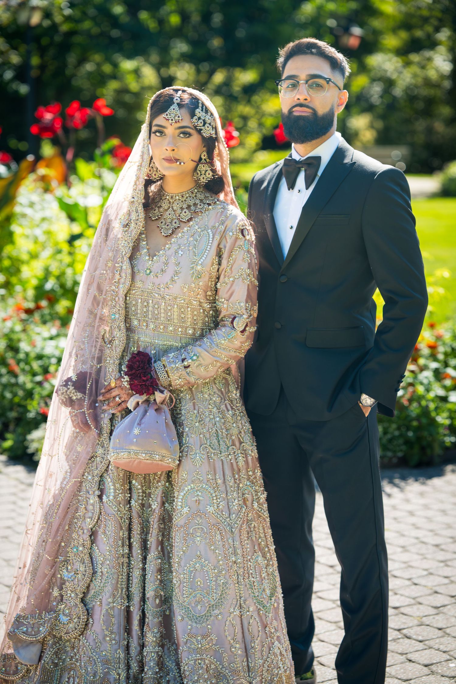 A wedding couple poses in a garden setting with the bride wearing an ornate beaded lehenga and the groom in a black tuxedo.