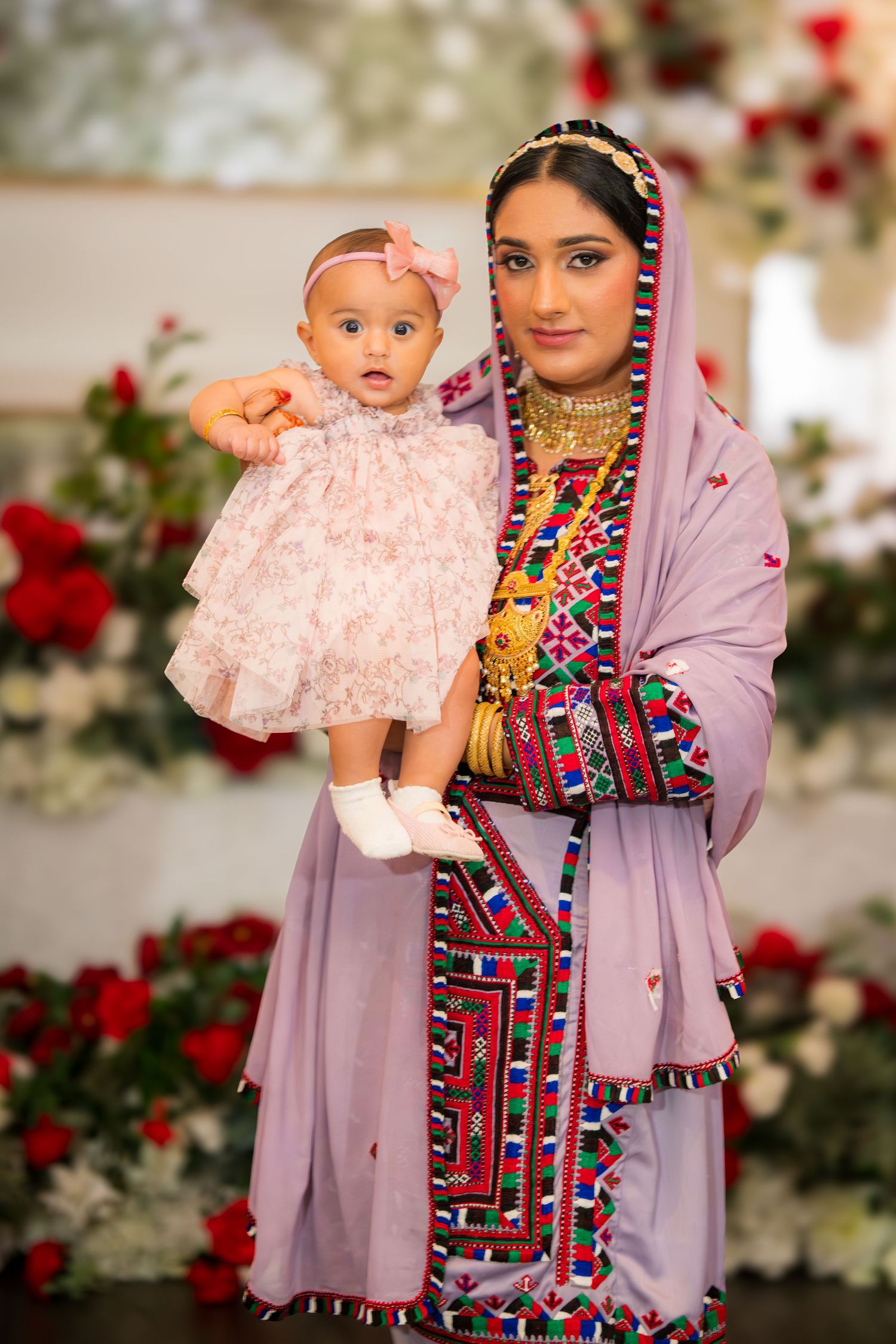 Traditional South Asian attire featuring ornate purple dress with colorful embellishments and holding baby in pink dress.