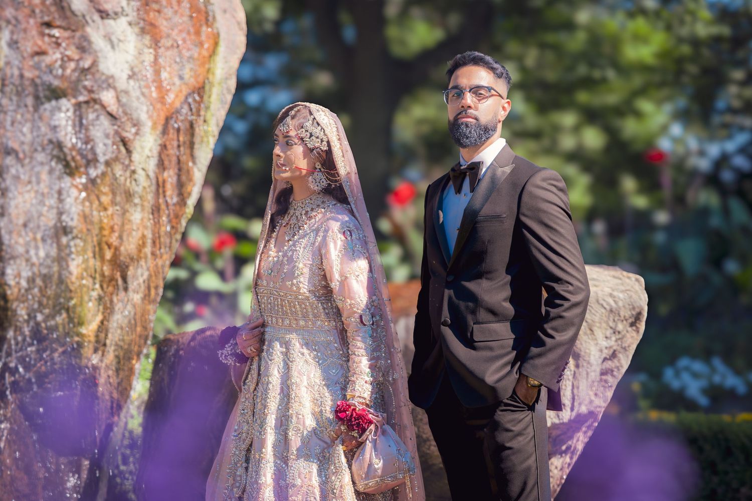 Wedding couple poses against blurred purple and natural outdoor background.