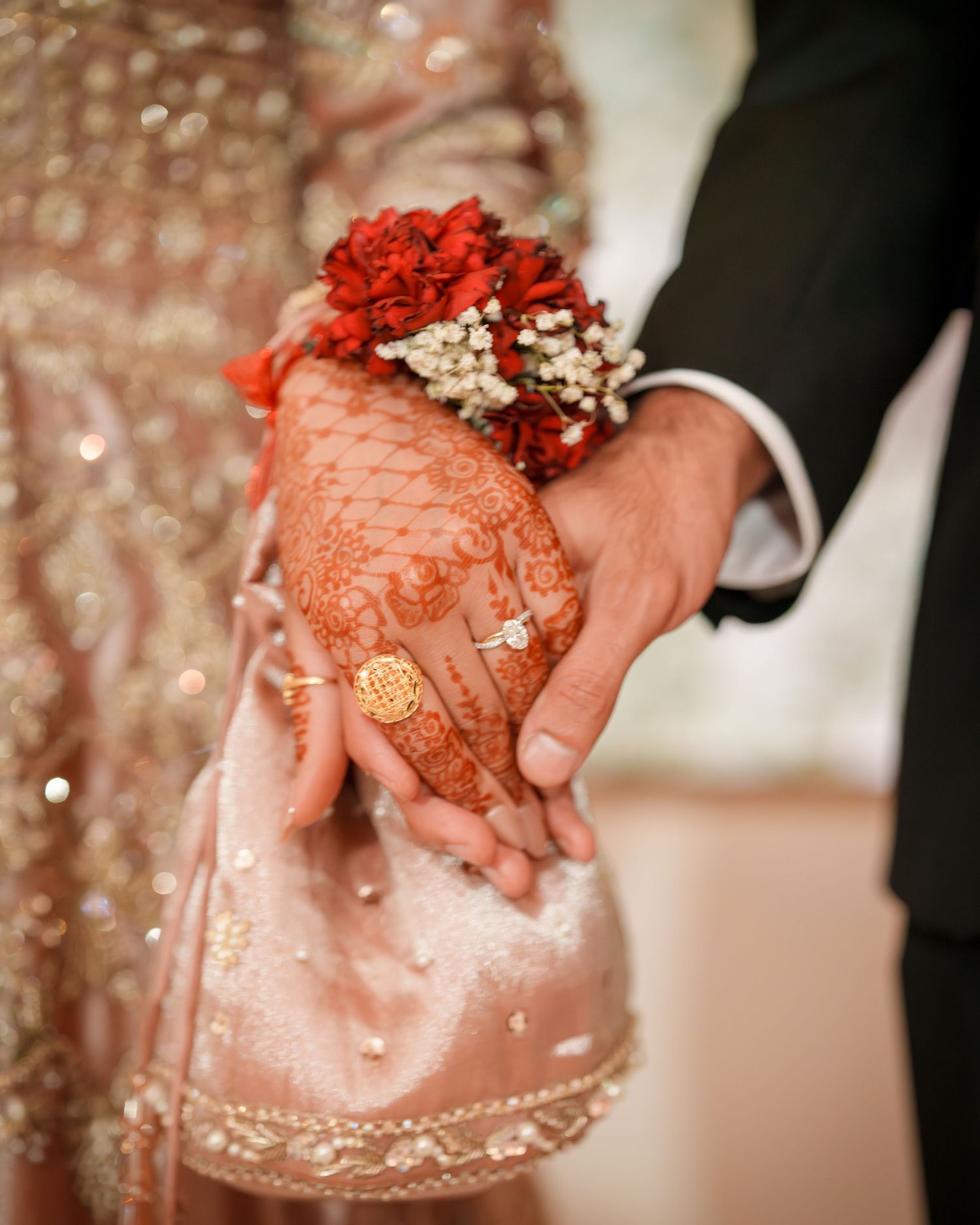 Close-up of hands with henna designs holding red flowers against an embellished pink dress.
