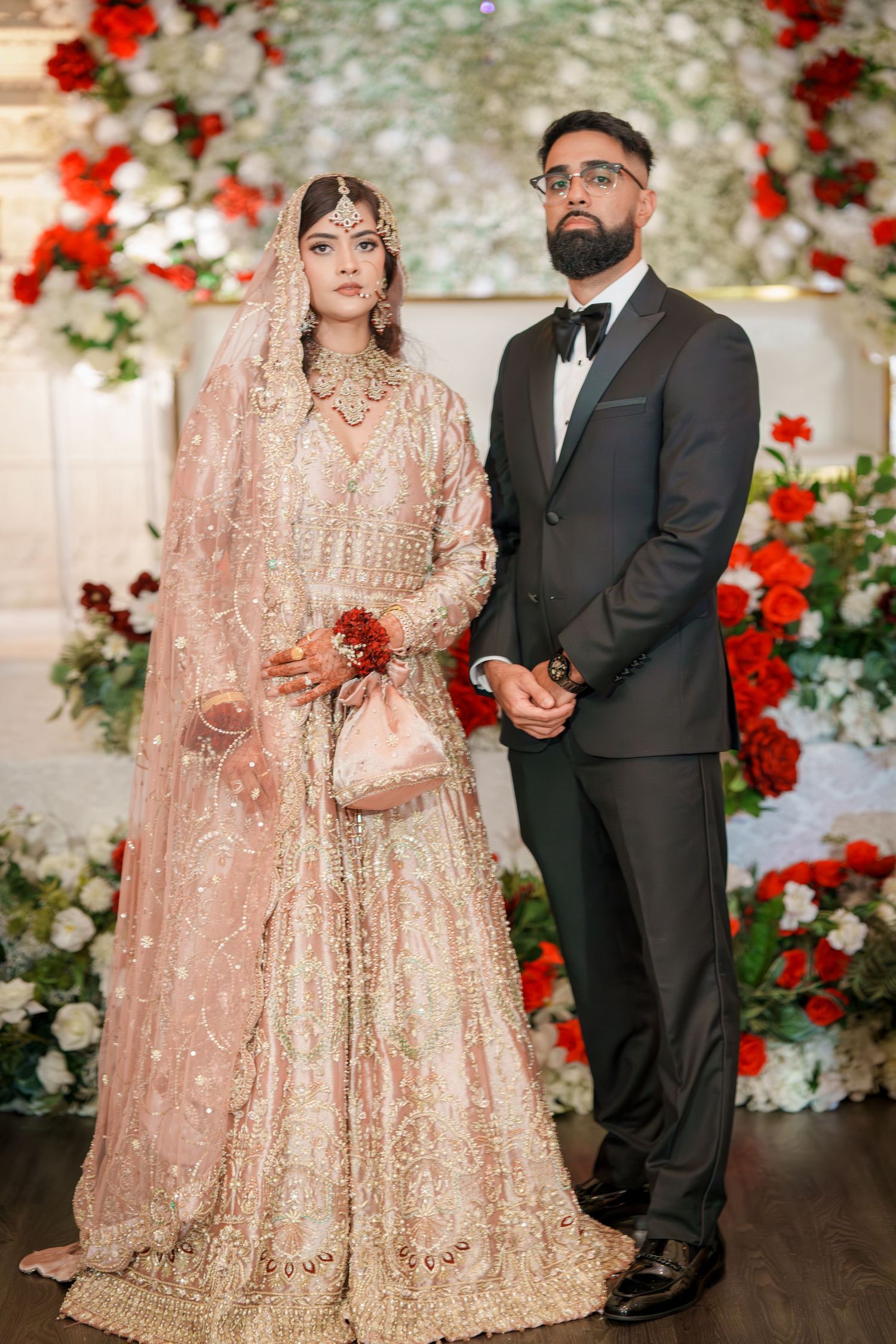 Wedding couple poses against floral backdrop with red and white roses wearing traditional South Asian attire.