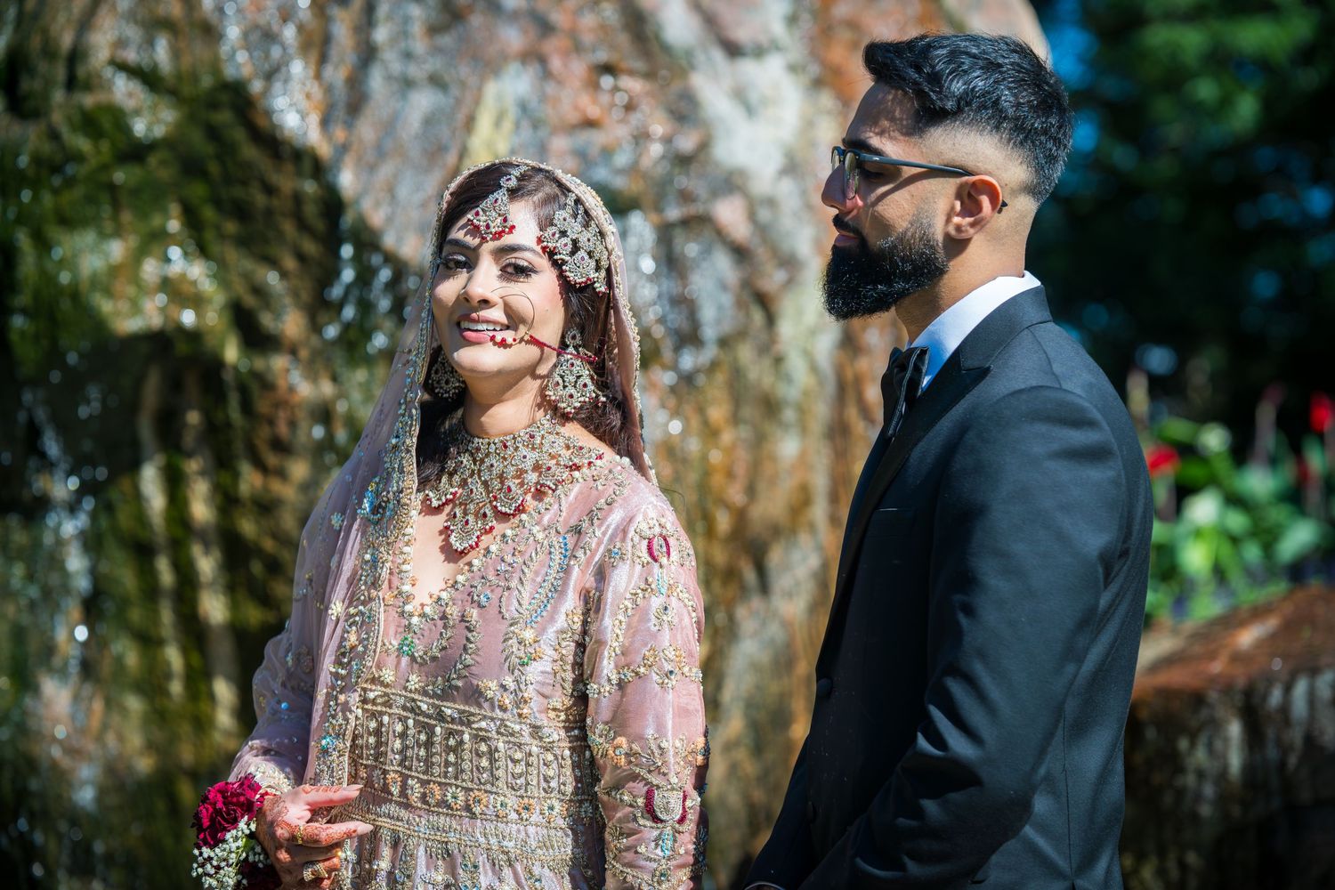 Traditional South Asian wedding couple standing before a rustic tree backdrop with ornate details.