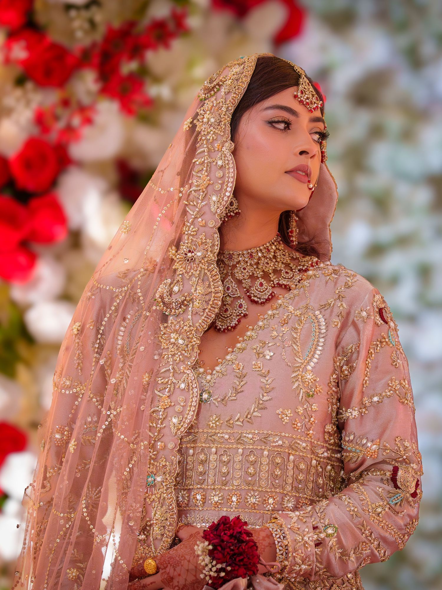 Bride in blush pink lehenga with intricate embroidery poses against red floral backdrop.