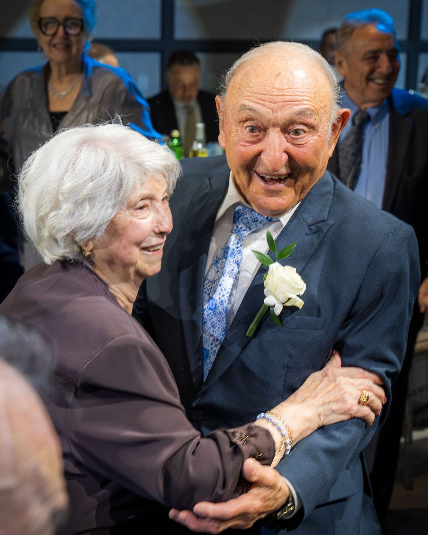 Elderly couple dancing joyfully at a celebration, man wearing boutonniere and blue tie, woman in brown top.