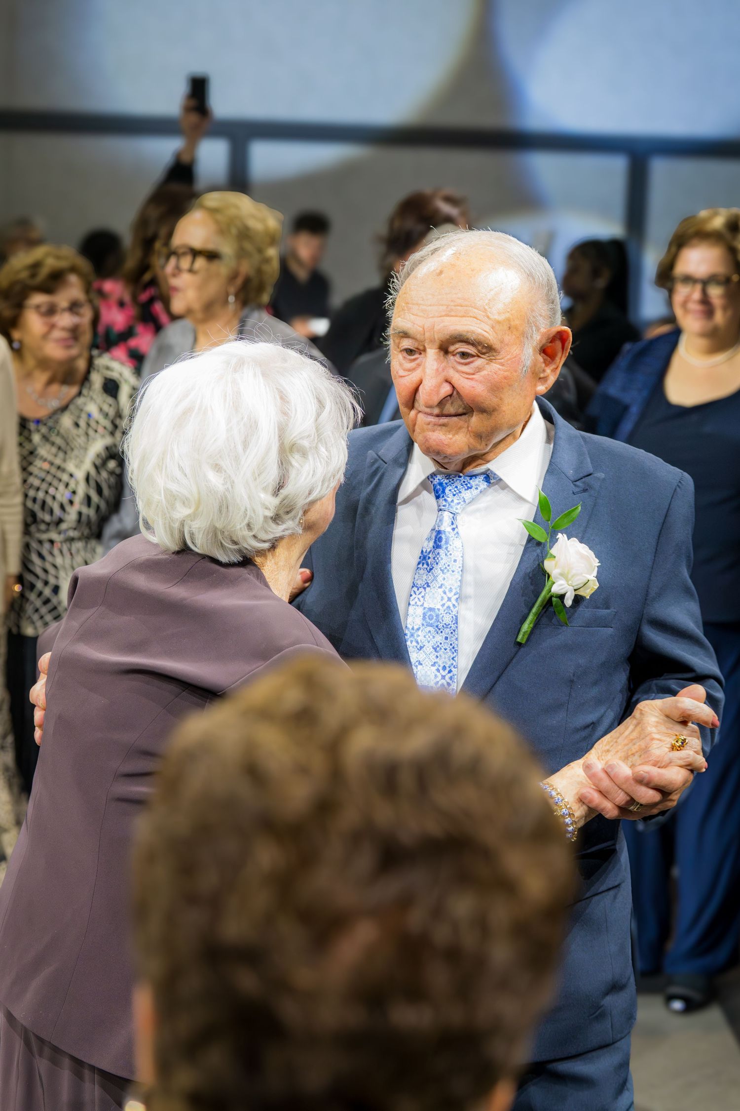 Elderly man in blue suit with boutonniere dancing with white-haired woman at a wedding anniversary celebration.