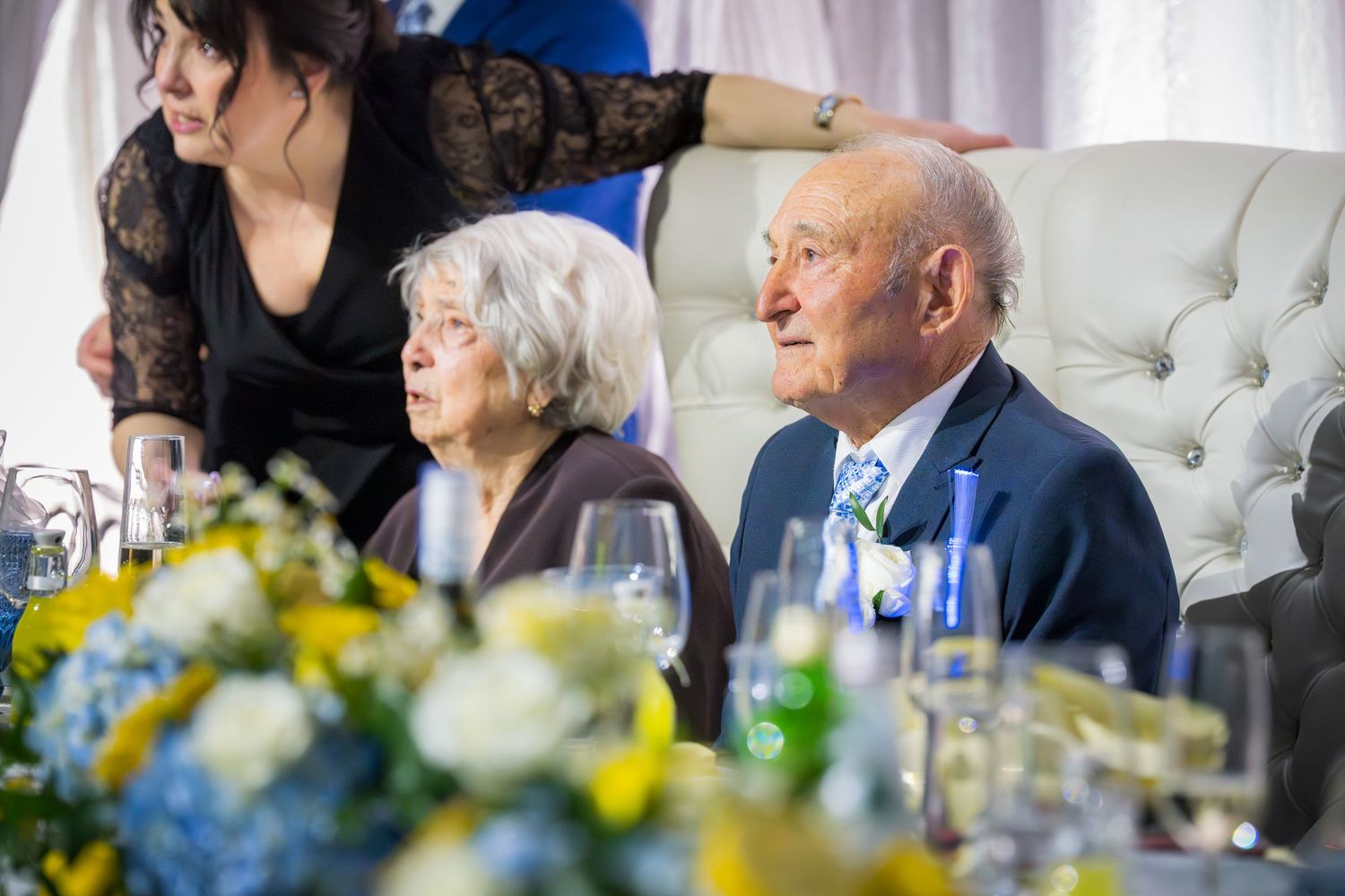 Senior couple seated at elegant wedding reception head table with blue and yellow floral decor.