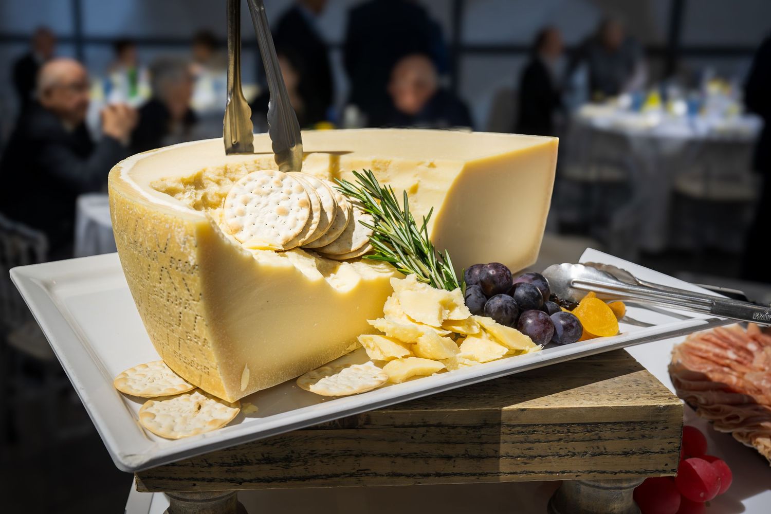 Large wheel of aged cheese displayed on wooden board with crackers, grapes, and rosemary at catered event.