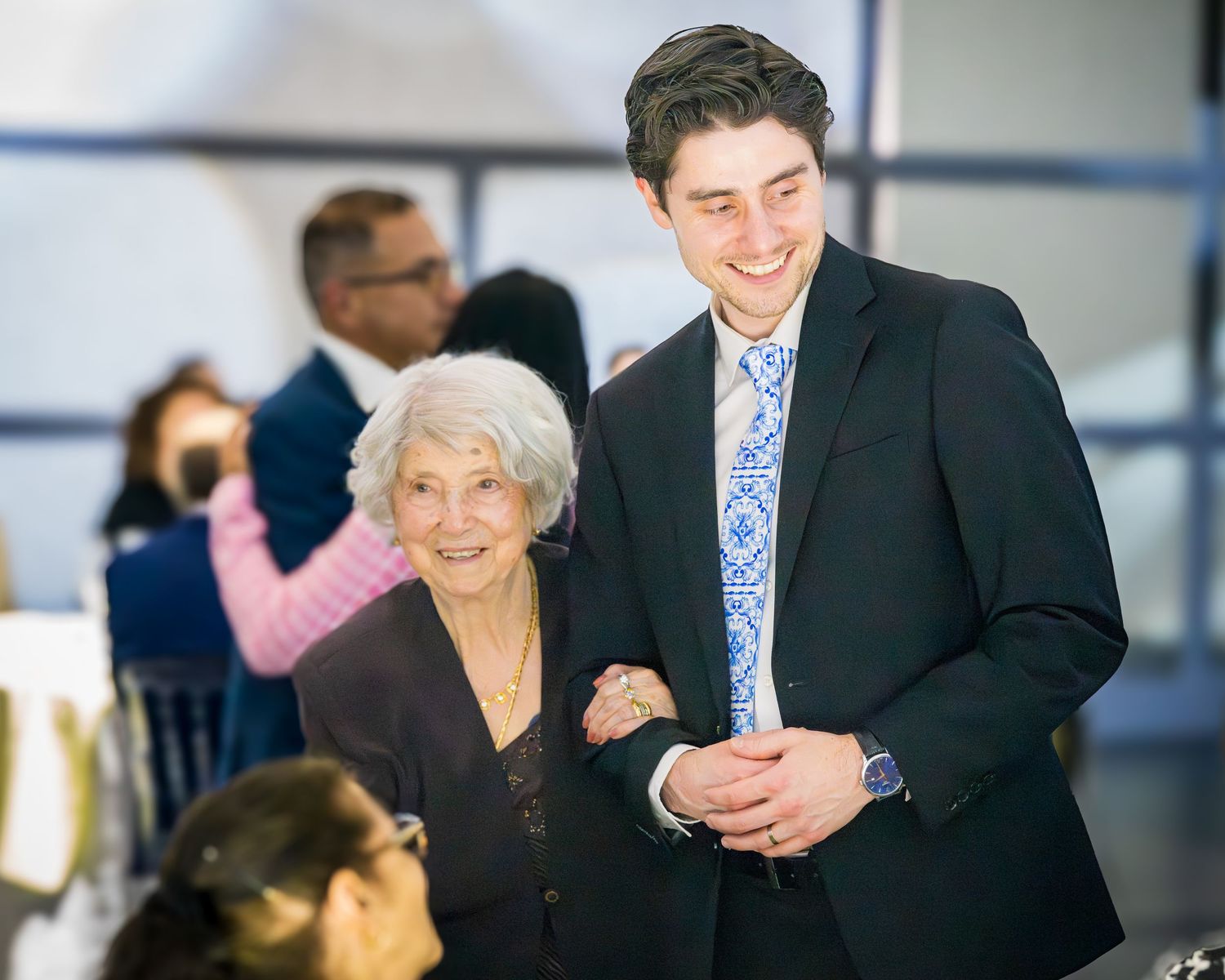 Elderly woman smiling and walking arm-in-arm with young man in suit at celebration.