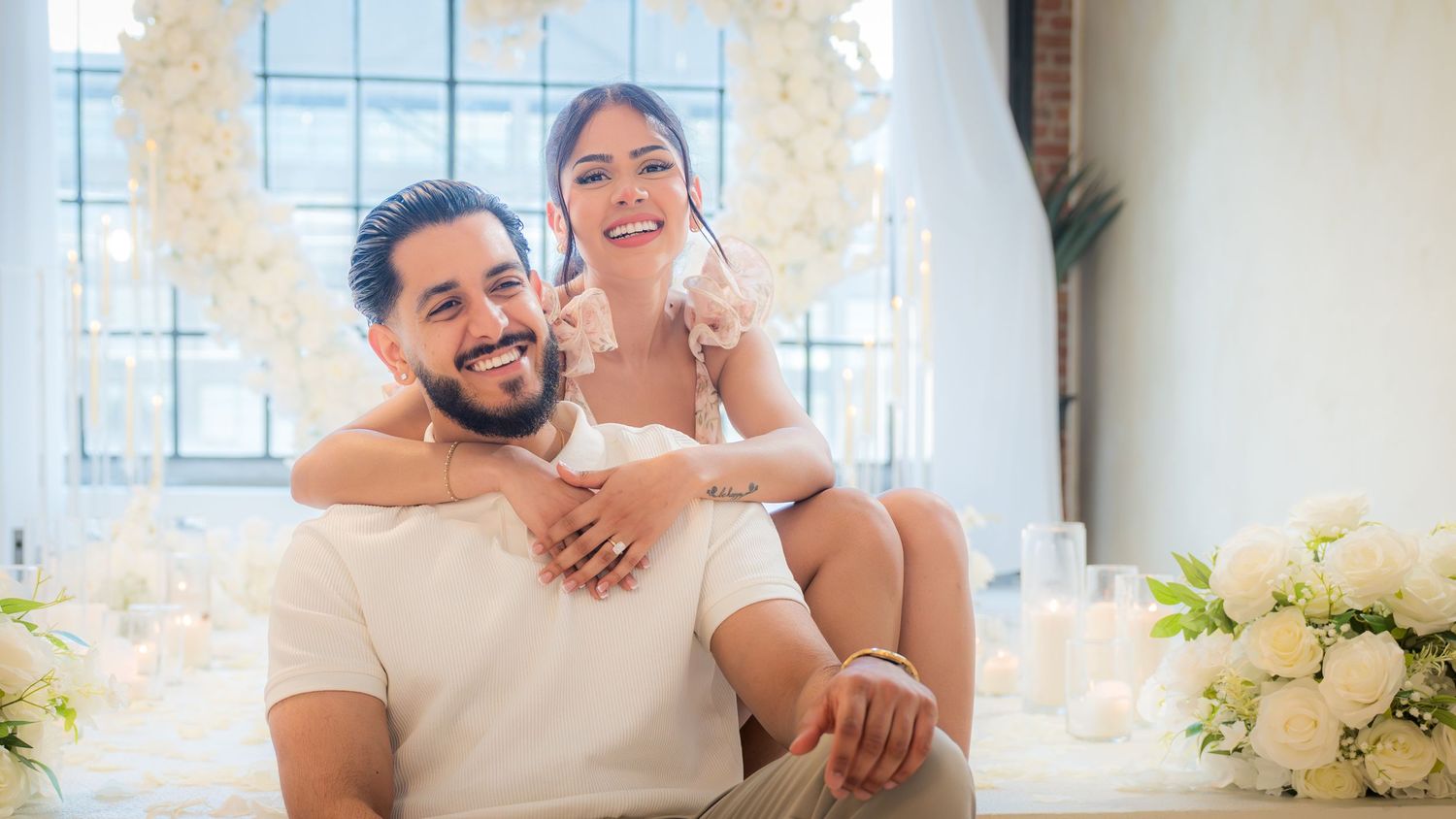Happy couple smiling together near white floral arch and candles in bright studio setting.