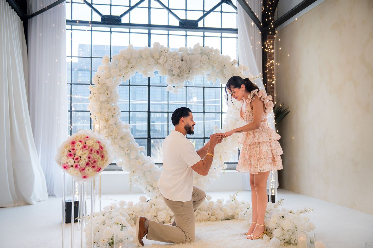 Man proposing on one knee to woman under white floral heart arch in elegant venue.
