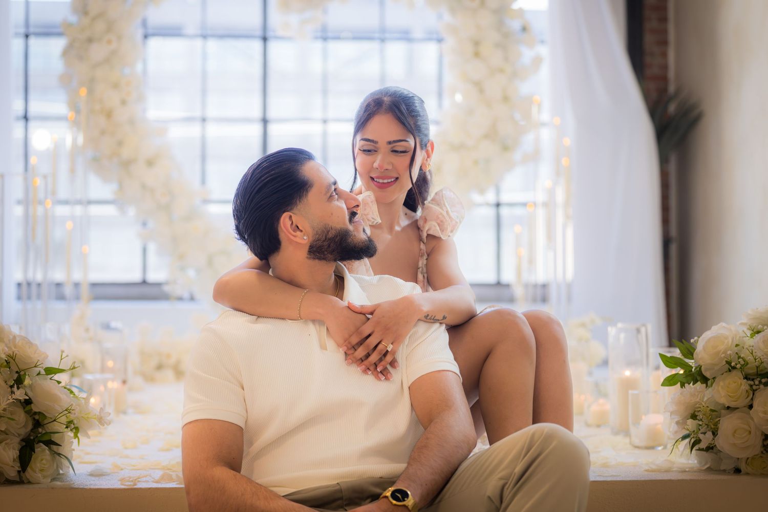 Couple sharing tender moment, woman sitting beside man in front of white floral heart arch backdrop.