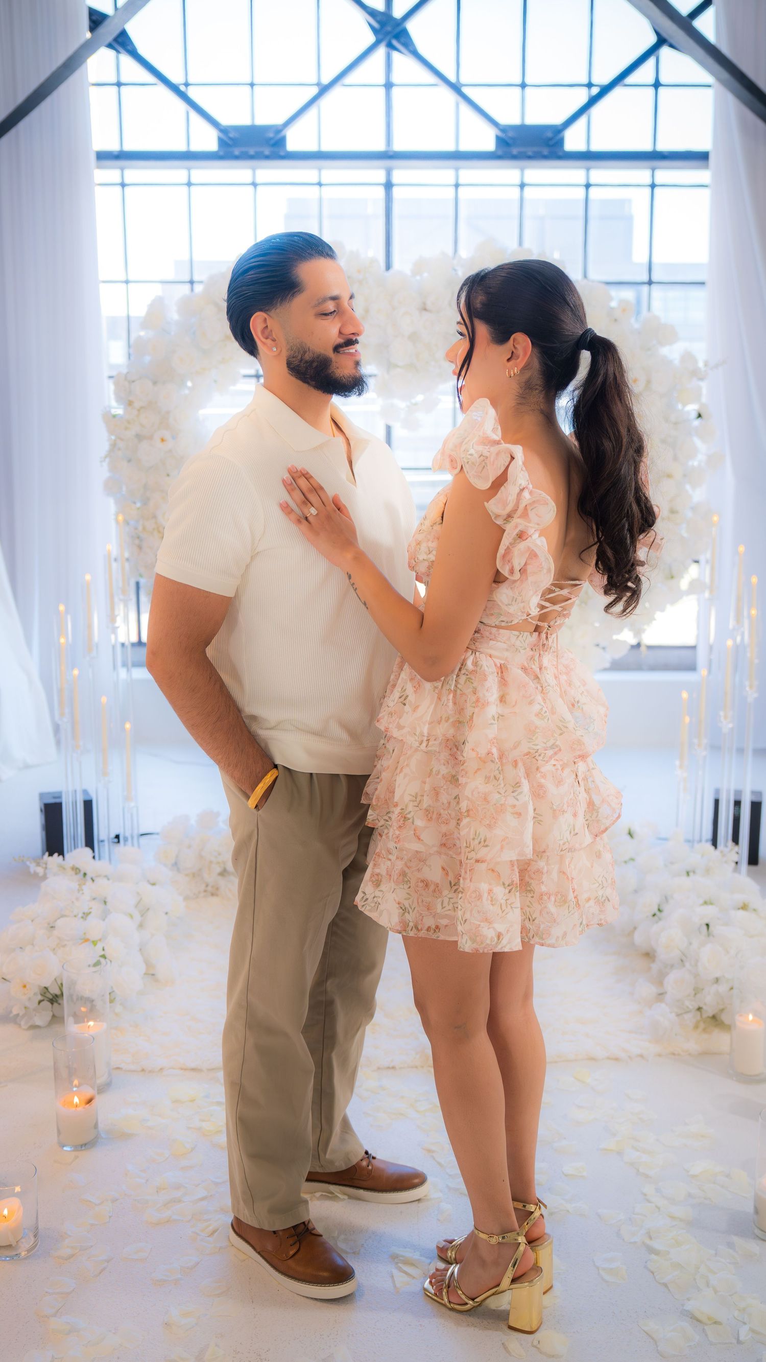 Couple gazing at each other in front of a white floral heart arch with candles during a romantic proposal.