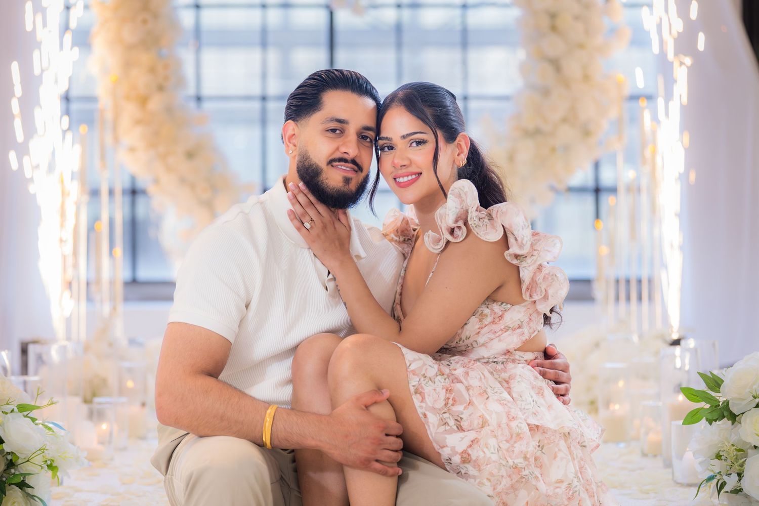 Newly engaged couple sitting together smiling in front of a white floral heart arch with candles.