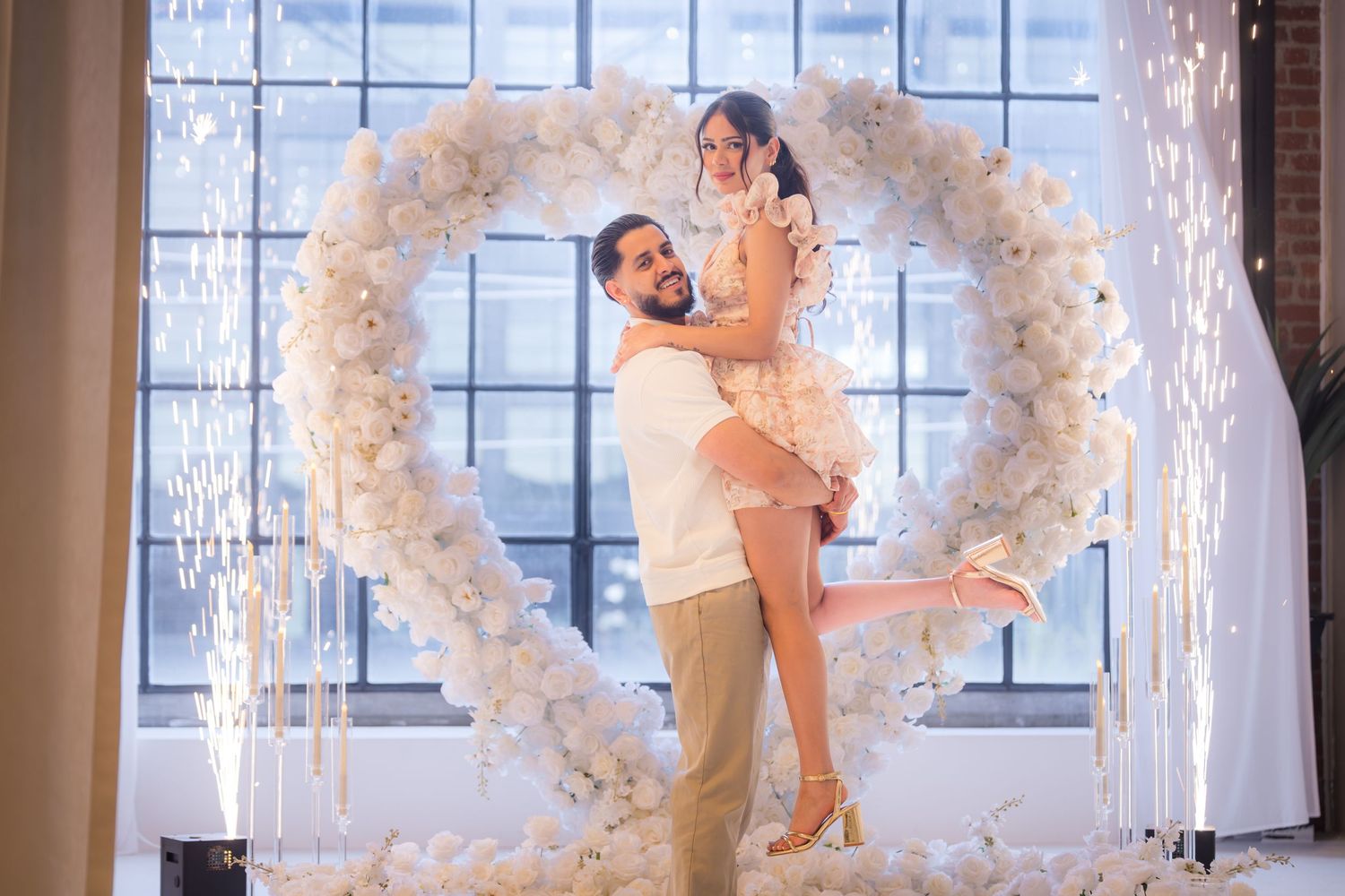 Man lifting woman in front of large white floral heart arch with sparkling lights backdrop.