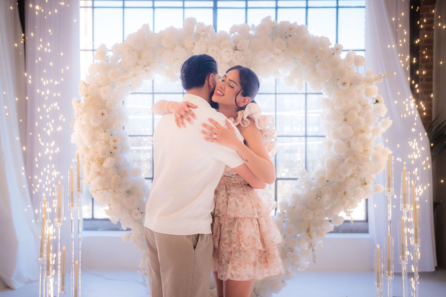 Couple sharing a romantic kiss under a large white floral heart arch with fairy lights at an elegant proposal setup.
