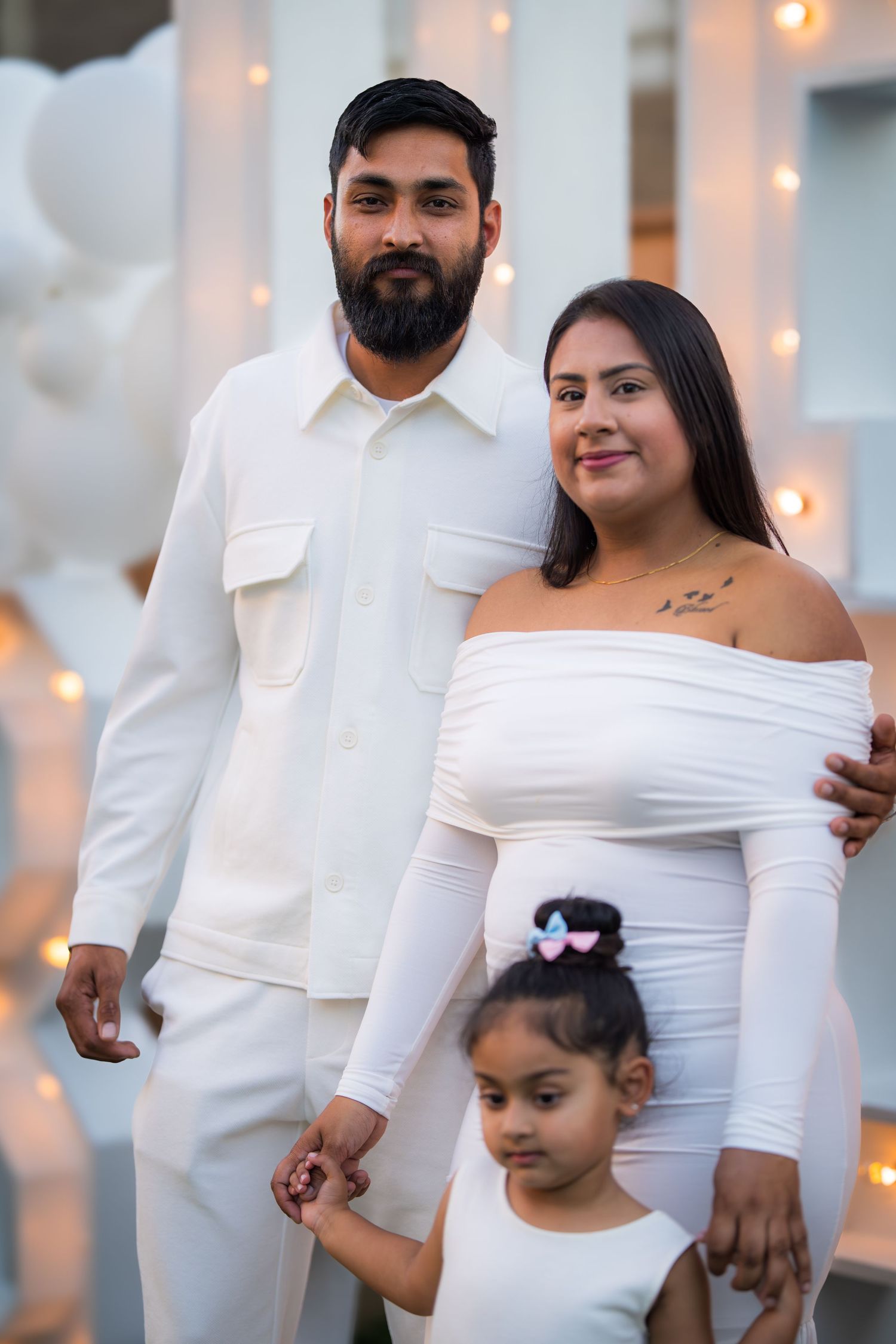A family of three in matching white outfits, with a bearded man, pregnant woman, and young girl posing together.