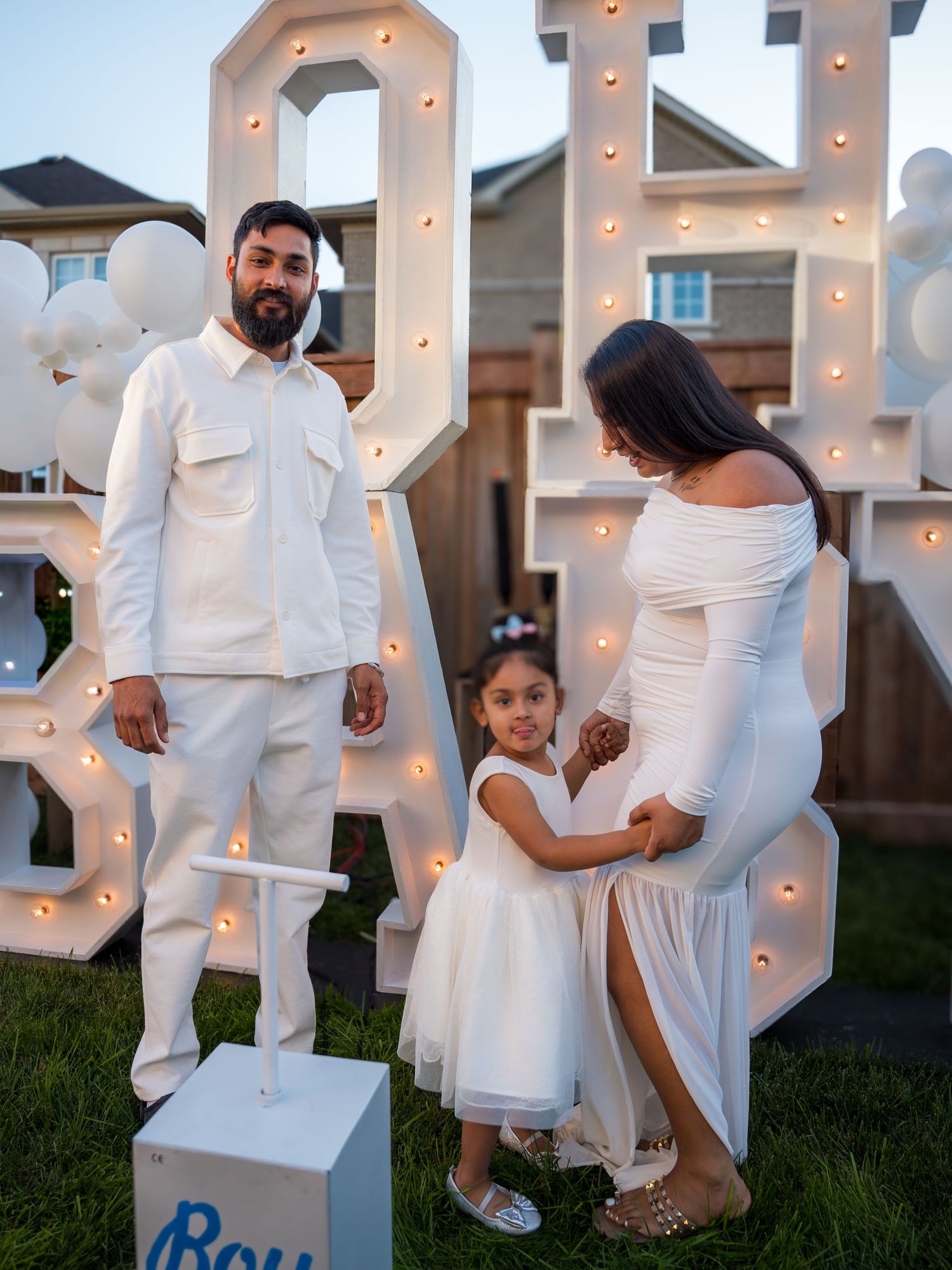 Family in white outfits posing in front of illuminated OH BABY marquee letters at a gender reveal party.