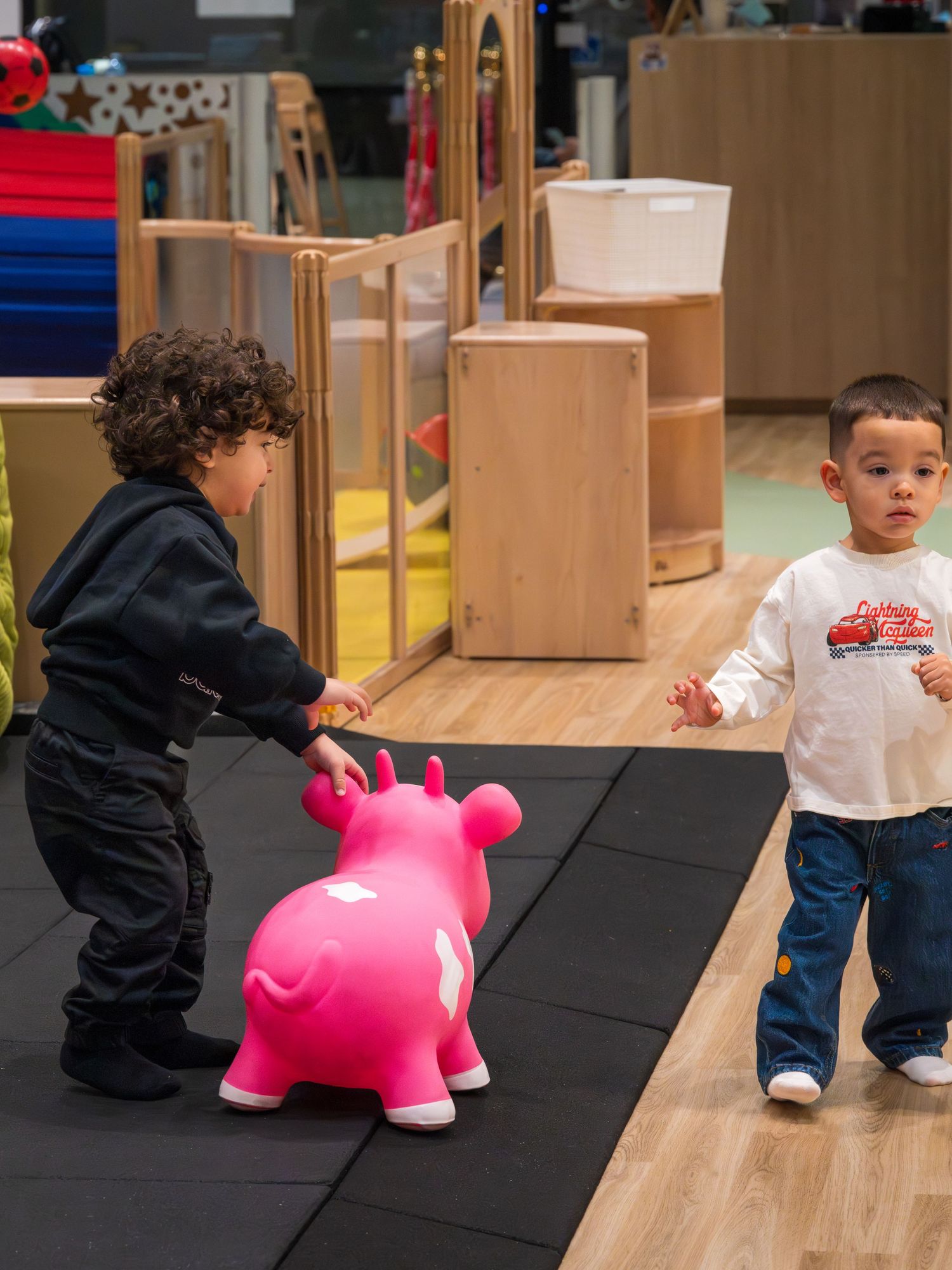 Two young children play with a pink bouncy toy horse in a colorful indoor play area.