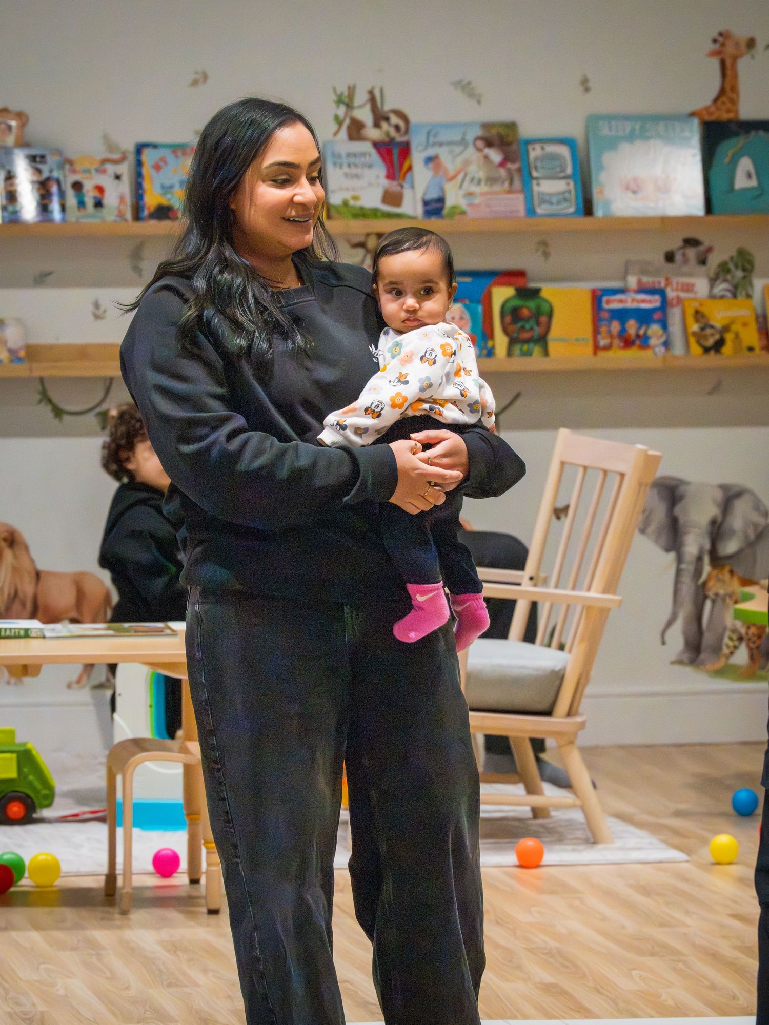 Woman in black outfit holding a baby in a floral onesie at a children's play library.