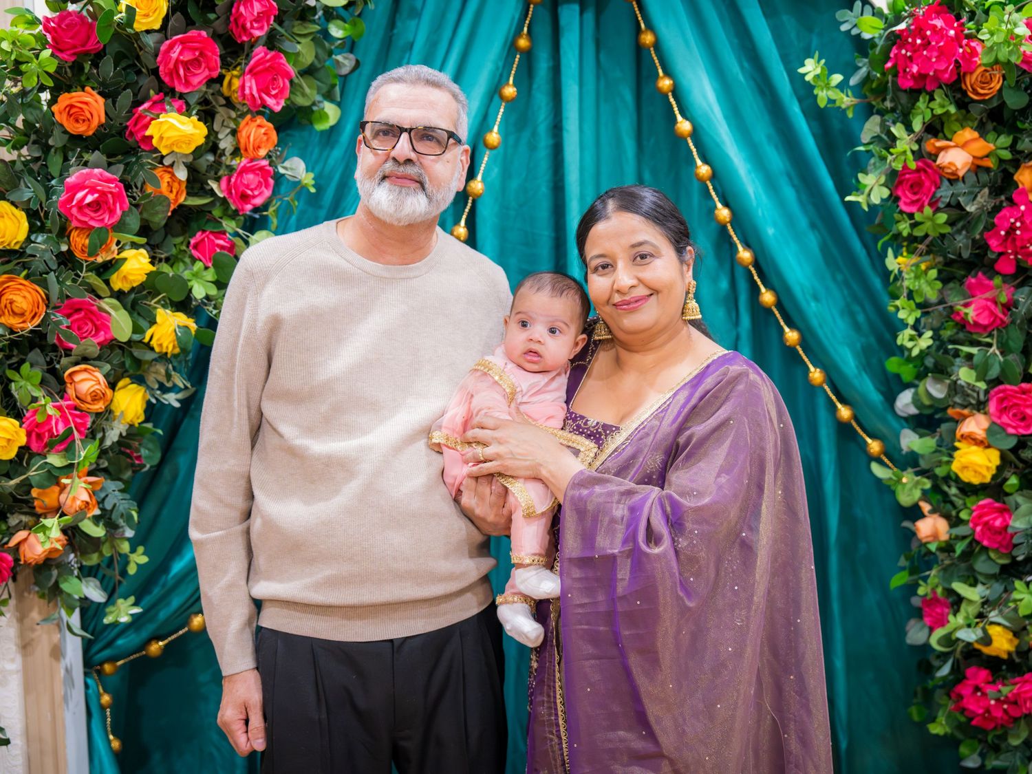 Grandparents in casual and purple sari holding baby before teal and floral ceremony backdrop.