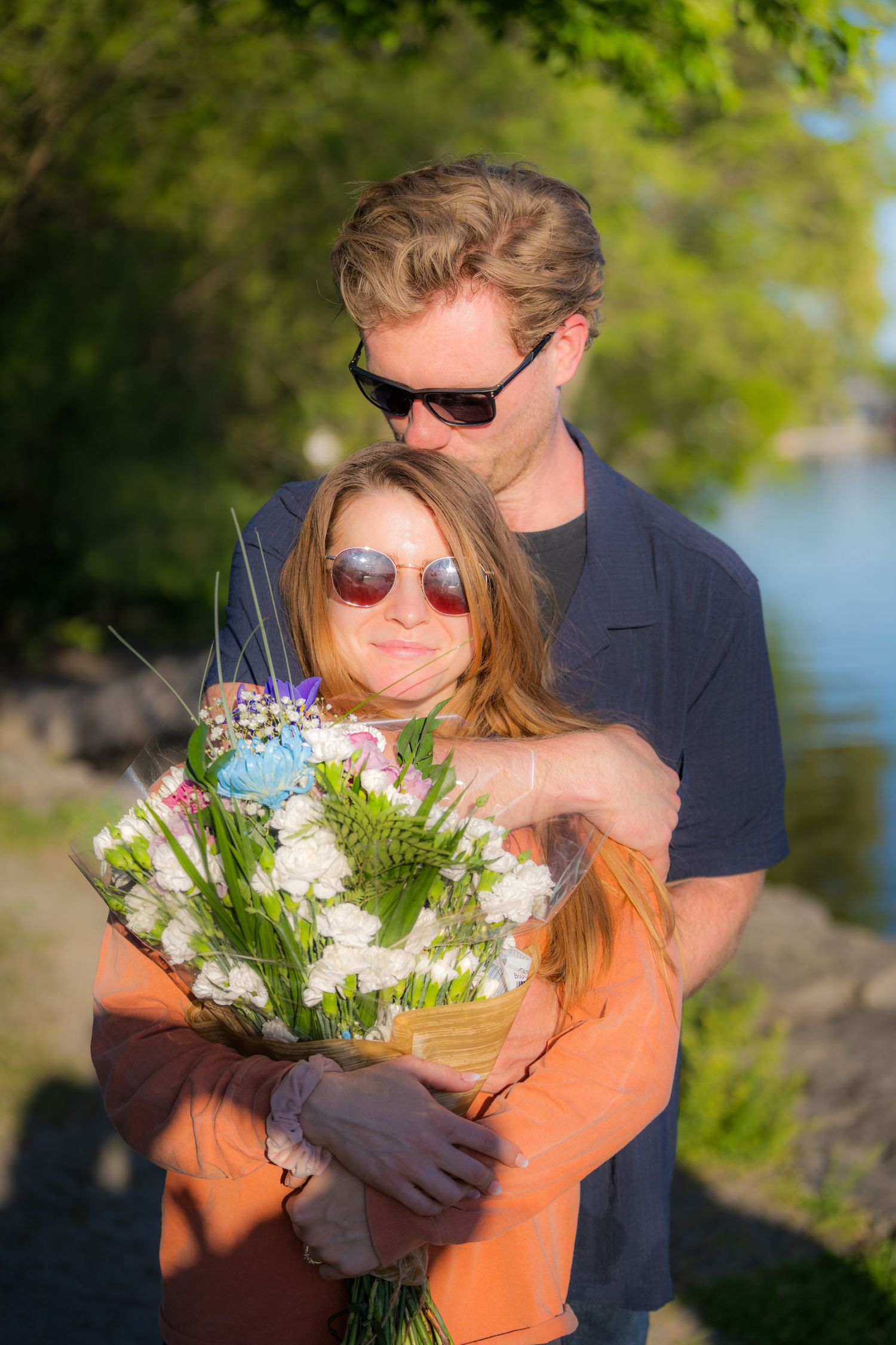 Man embracing woman from behind as she holds a large colorful bouquet near a sunny lakeside.