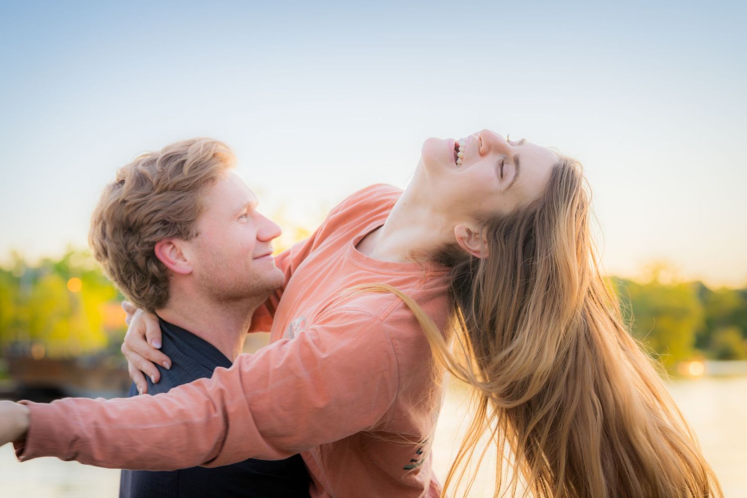 Happy couple laughing and embracing outdoors at sunset, woman leaning back joyfully with long flowing hair.