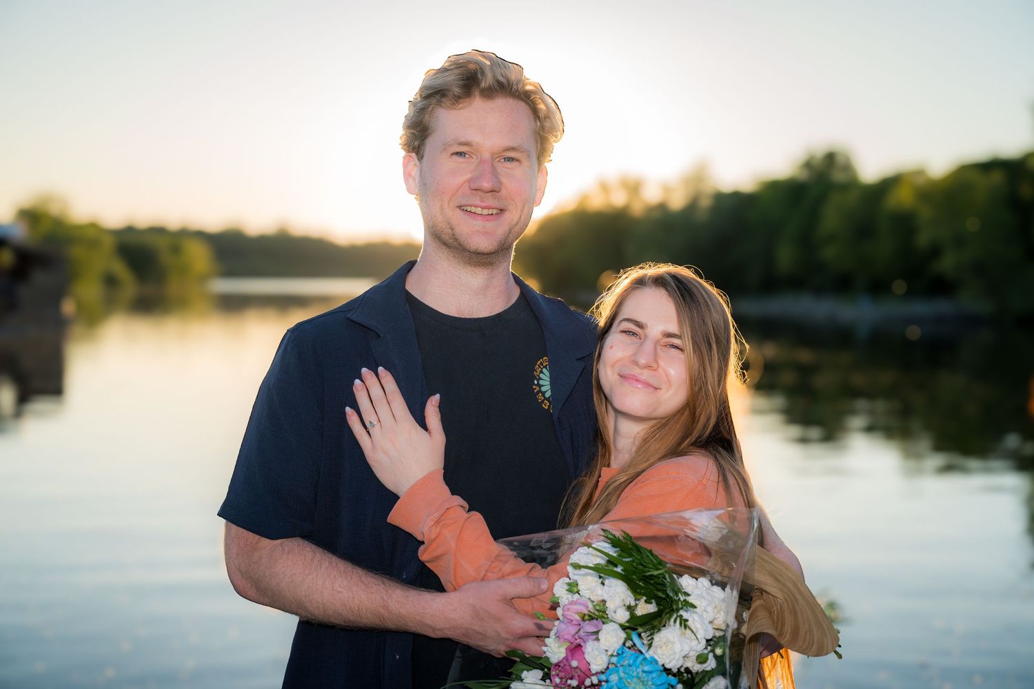 Happy couple posing near a lake at sunset, woman holding a bouquet of flowers after a proposal.