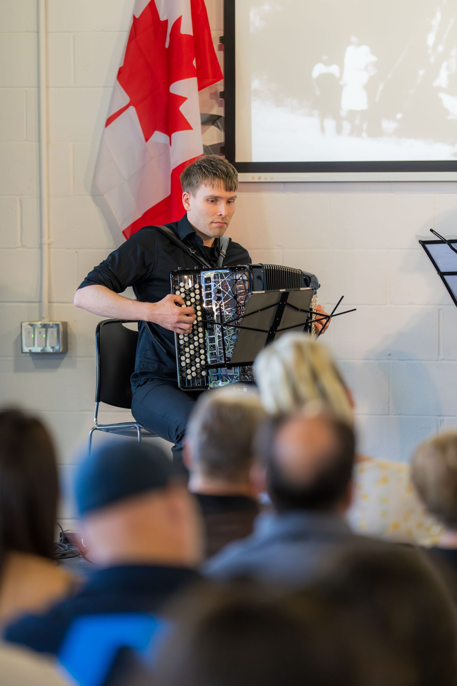 Young man playing accordion at Canadian cultural event with maple leaf flag displayed behind him.