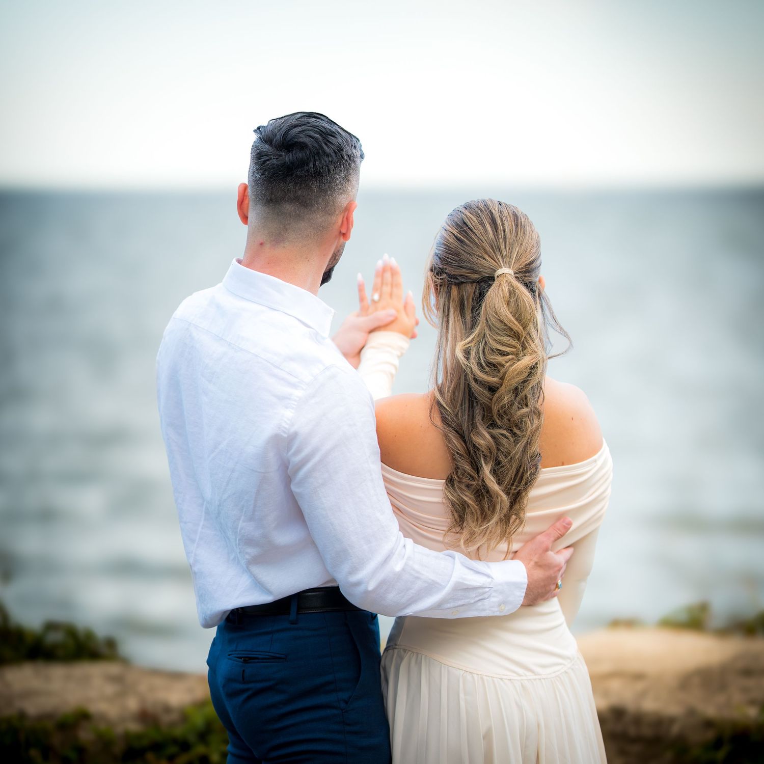 A couple in formal attire poses together near the ocean.